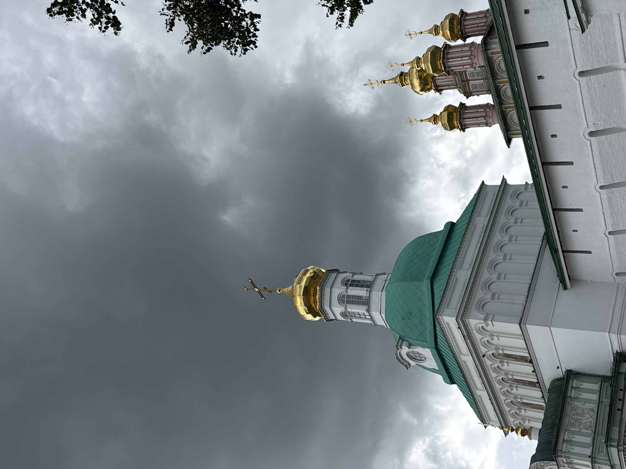 This is a medium-angle, outdoor, vertical shot of the Trinity Lavra of St. Sergius in Sergiyev Posad, Russia. The image captures several of the monastery's domes and a section of its white wall. Dominating the upper left is a large, bright green domed structure with white architectural accents and arched windows. Above and to the right of this green dome, a smaller, gold-colored dome topped with a cross stands out against a dramatic, dark gray, cloudy sky. In the middle ground to the right, a cluster of smaller, ornate gold domes, also topped with crosses, are visible, some with pink architectural details. A section of a long, white wall with a green roofline runs across the bottom of the frame, forming a lower boundary of the scene. Dark green foliage from trees is visible at the top right of the image. The overall lighting is dim, suggesting an overcast day, possibly before or during a storm, due to the heavy, dark clouds. There are no people or animals visible in the frame.