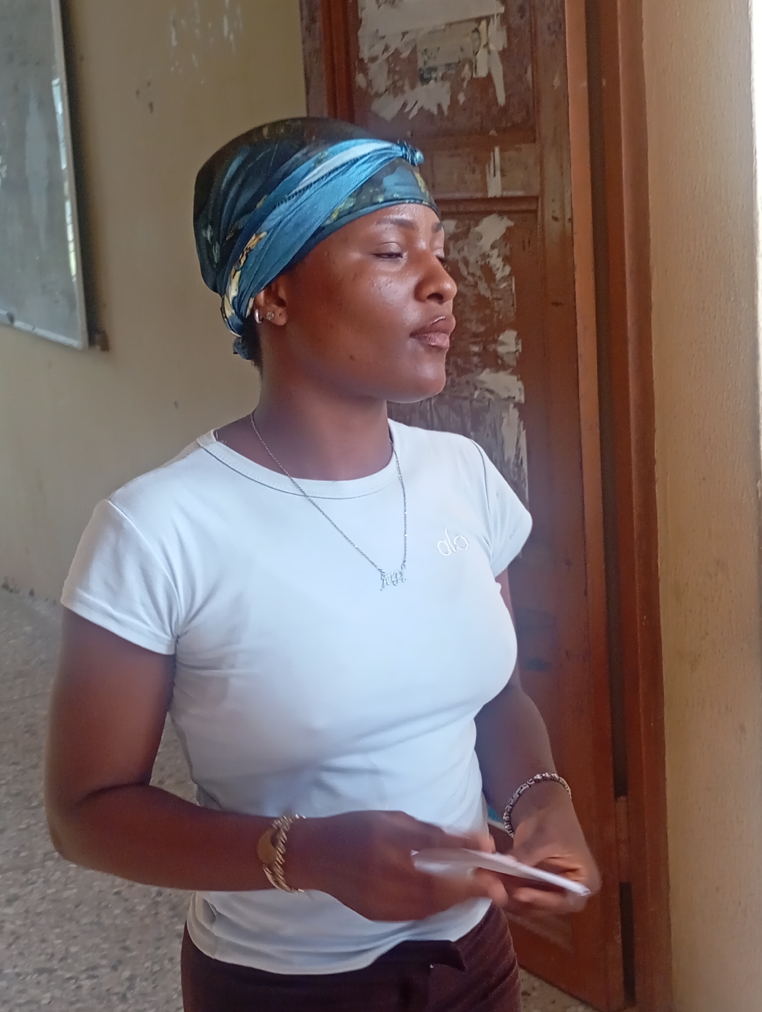 A woman stands indoors, possibly in Ukwu-Abwa, Nigeria, although the location cannot be definitively confirmed from the image alone. She wears a blue headscarf, a white t-shirt with the word "alo" printed on it, a silver necklace, and a brown skirt. She also has jewelry on her wrists. She holds a white object in her hands. Behind her is a door with peeling paint and an off-white wall. A board is visible on the wall to the left. The floor appears to be tiled.
