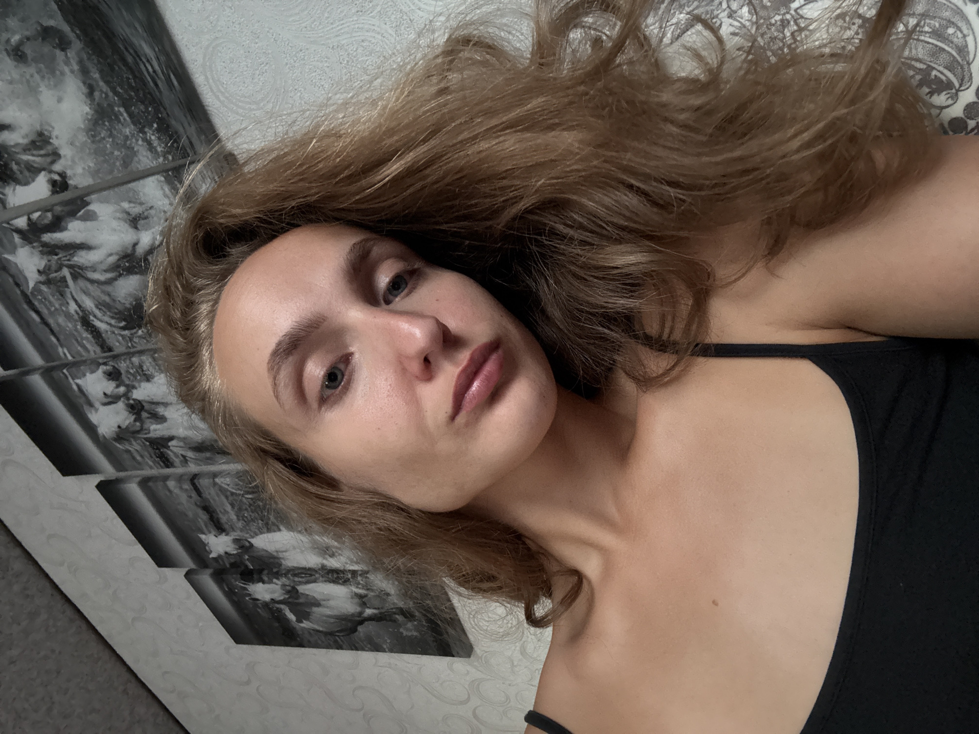 A young woman with long, wavy blonde hair is shown in a close-up selfie. She is wearing a black tank top. The background consists of a textured wallpaper and a four-panel wall art depicting horses. The scene appears to be indoors, possibly in a residential setting. There is no visible indication of the city of Kuyeda, Russia.