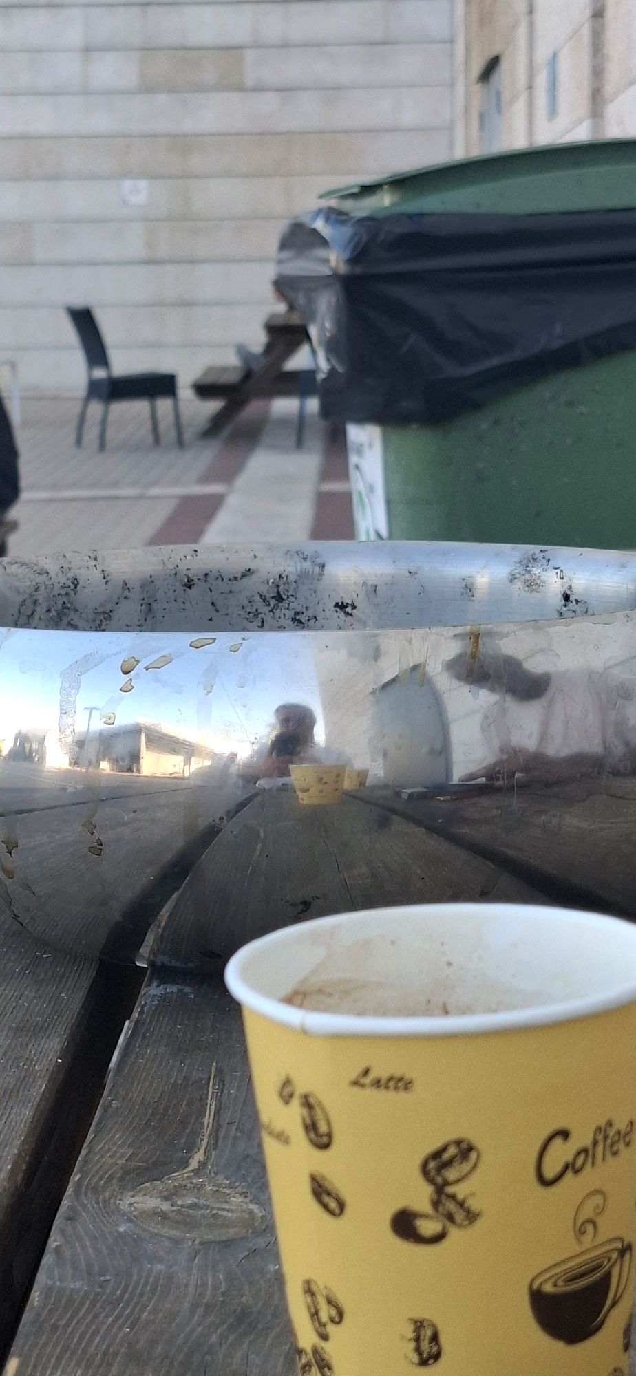 In Kiryat Gat, Israel, this image captures a close-up view of a yellow paper cup of coffee resting on a weathered wooden picnic table. The cup, adorned with coffee bean illustrations and the words "Latte" and "Coffee," is partially filled. The table's surface shows distinct wood grain and knots. In the background, a metallic railing, likely part of an outdoor seating area, reflects the surroundings. The reflection reveals a person wearing a white shirt, seated at the table, with two small yellow bowls in front of them. Further in the background, a green trash bin with a black bagliner is visible, along with a blurred image of a building with horizontal light-colored brickwork and a dark-colored chair. The scene appears to be an outdoor public space, possibly a park or a seating area outside a cafe or municipal building.