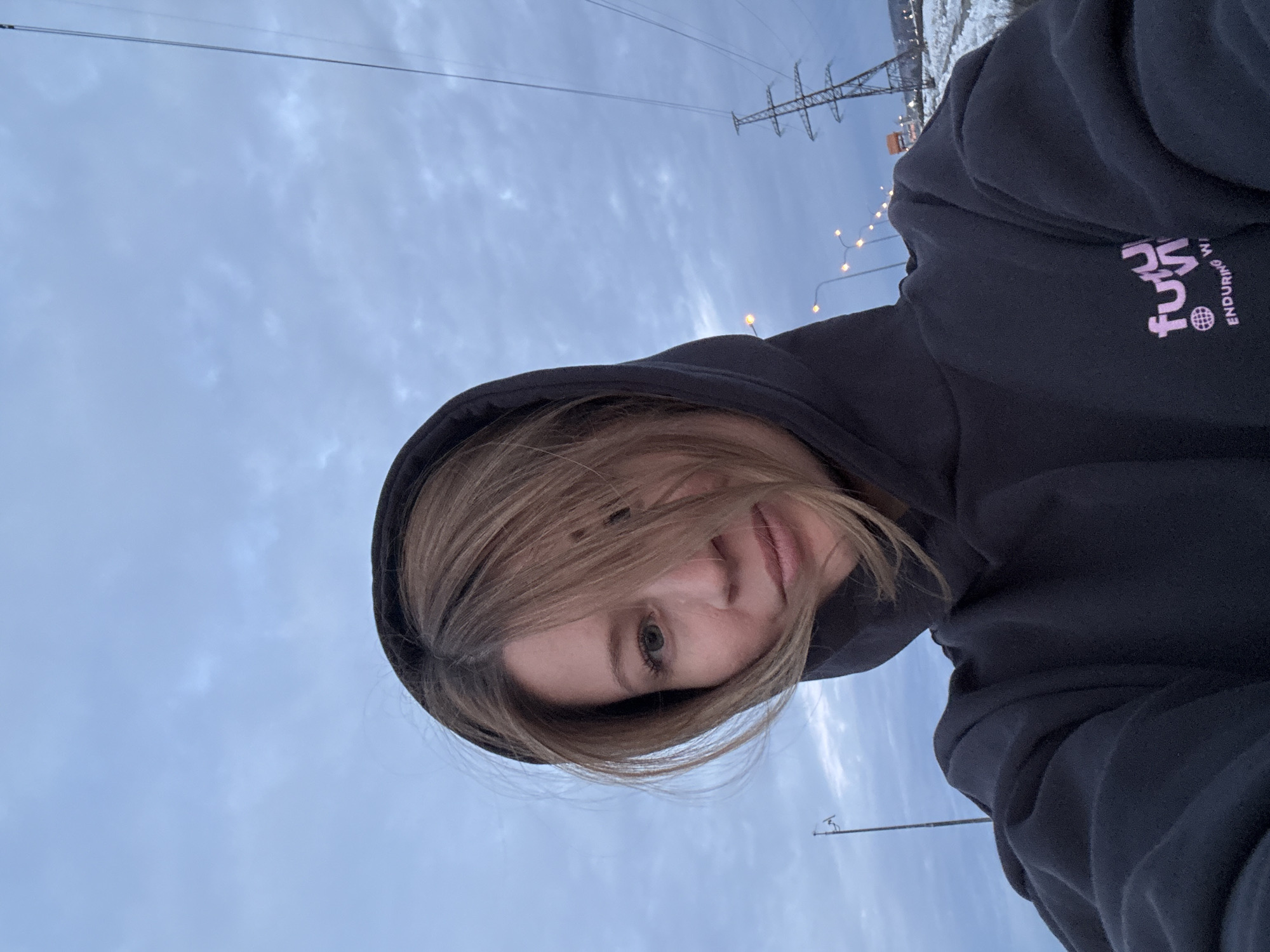 An individual, appearing female, is depicted in a close-up selfie against an outdoor backdrop in Chaikovsky, Russia. She wears a dark hooded sweatshirt with the hood up, framing her face; light brown hair is partially windblown across her left eye and forehead. A pink graphic featuring stylized text, potentially "futu" or "future", with a globe icon and the partial phrase "ENDURING W", is visible on the sweatshirt's chest. The sky is overcast with a blue-grey hue, suggesting dim light conditions. In the background, illuminated streetlights are visible to the left, and a large metal lattice-type electricity pylon stands prominently to the right. Patches of snow are present on the ground beneath the pylon.