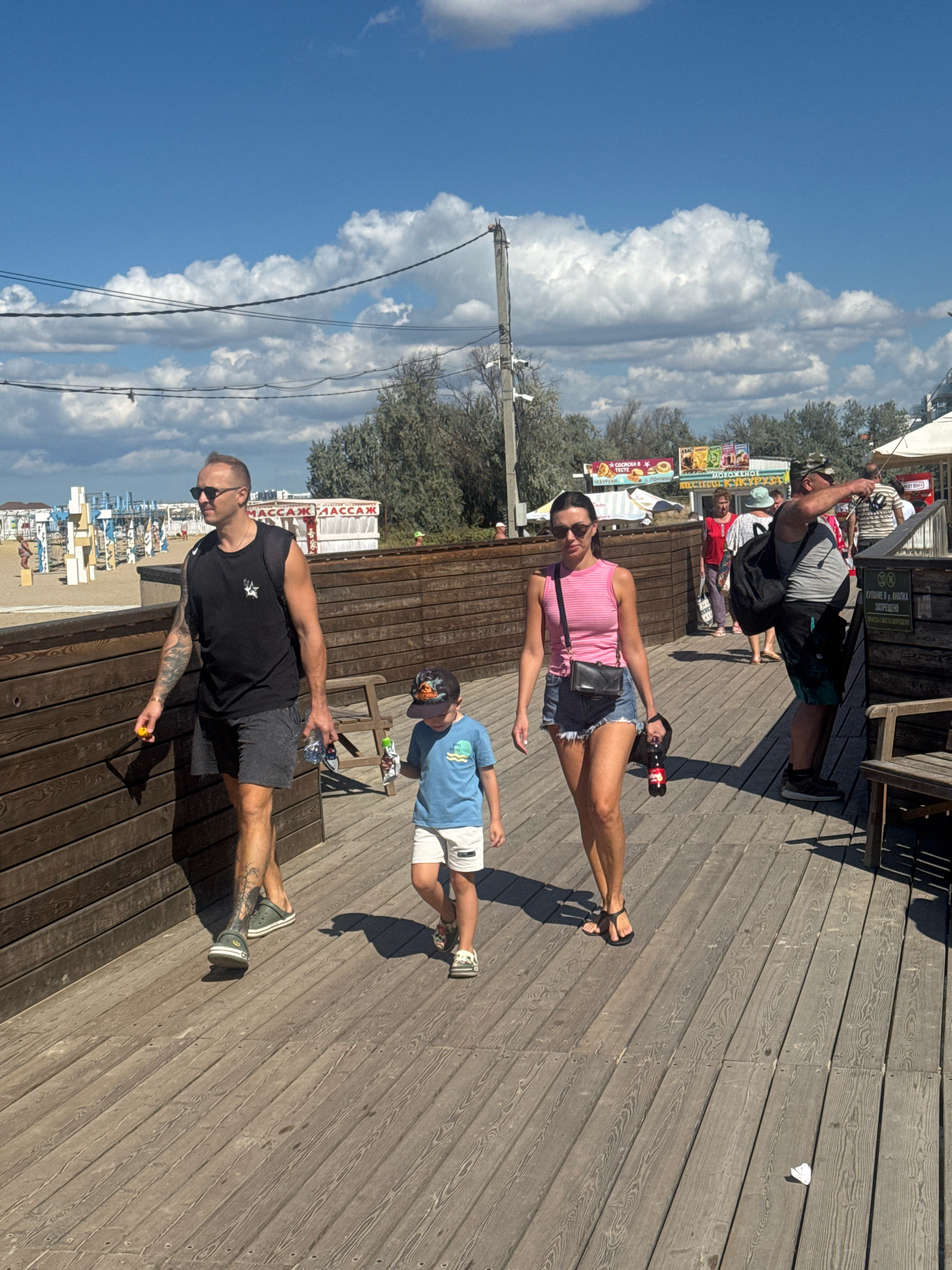 In this sunny outdoor scene, a family walks along a wooden boardwalk. A man in sunglasses and a black sleeveless shirt walks with a bottle of water and a yellow object. Beside him, a young boy in a blue t-shirt and white shorts holds a toy. Behind them, a woman in a pink tank top and denim shorts walks with a black purse and a bottle of soda. In the background, a man in a camouflage hat points towards something off-camera. The boardwalk is lined with wooden fences and leads towards a beach with lounge chairs and umbrellas. Several food stalls are visible, with signs displaying text in Cyrillic, including "МАССАЖ" (massage), "МОРОЖЕНОЕ" (ice cream), and "ЧЕБУРЕКИ" (a type of pastry). The sky is blue with fluffy white clouds, suggesting a pleasant, warm day. The overall atmosphere is casual and recreational, typical of a beach resort.