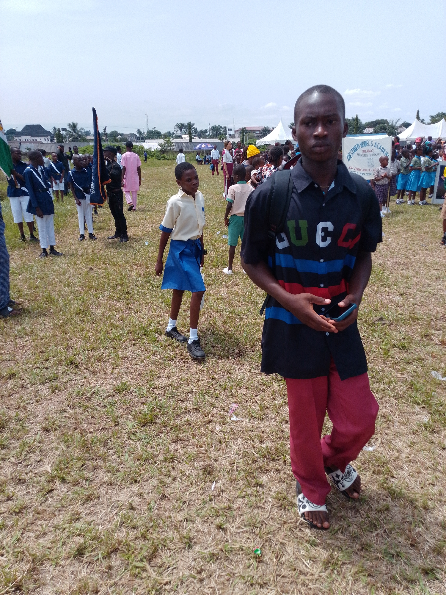 The scene depicts an outdoor event in Abak, Nigeria, likely a school function, taking place on a grassy field under a partly cloudy sky.
A young man stands prominently in the foreground. He wears a black shirt with the word "GUCC" in large, colored letters across the upper part, red and blue stripes underneath, and red trousers with patterned sandals. He carries a backpack and holds a smartphone in his hands.
Behind him, a young girl in a school uniform, consisting of a beige shirt and blue skirt, walks in the same direction. To the left of her, a group of students dressed in blue and white uniforms are lined up, some holding flags.
In the background, a crowd of people is visible, along with tents and other temporary structures, suggesting an organized event. A banner is partly visible near the crowd that appears to say “Destined Nobels Academy” suggesting a school function of some kind. The environment is open and expansive, with scattered trees in the distance and a few buildings visible beyond the crowd.