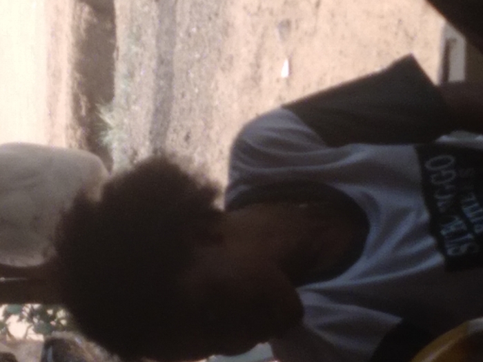 The image displays a close-up of an individual located in Agege, Nigeria, viewed from the chest up. The person has dark, curly hair, with their face obscured by shadow and blur. They are wearing a short-sleeved t-shirt; the main body is light-colored, and the sleeves are dark. The word "STRONGGO" is partially visible in dark lettering on the front of the shirt. A thin, light-colored chain or necklace is present around the individual's neck. The person is looking downwards and towards the left of the frame. The background consists of reddish-brown dirt ground with sparse patches of green vegetation. An indistinct, large, light-colored, rounded object is visible in the upper right background, and a dark, vertical element is partially visible on the upper left, partially obscured by foliage.