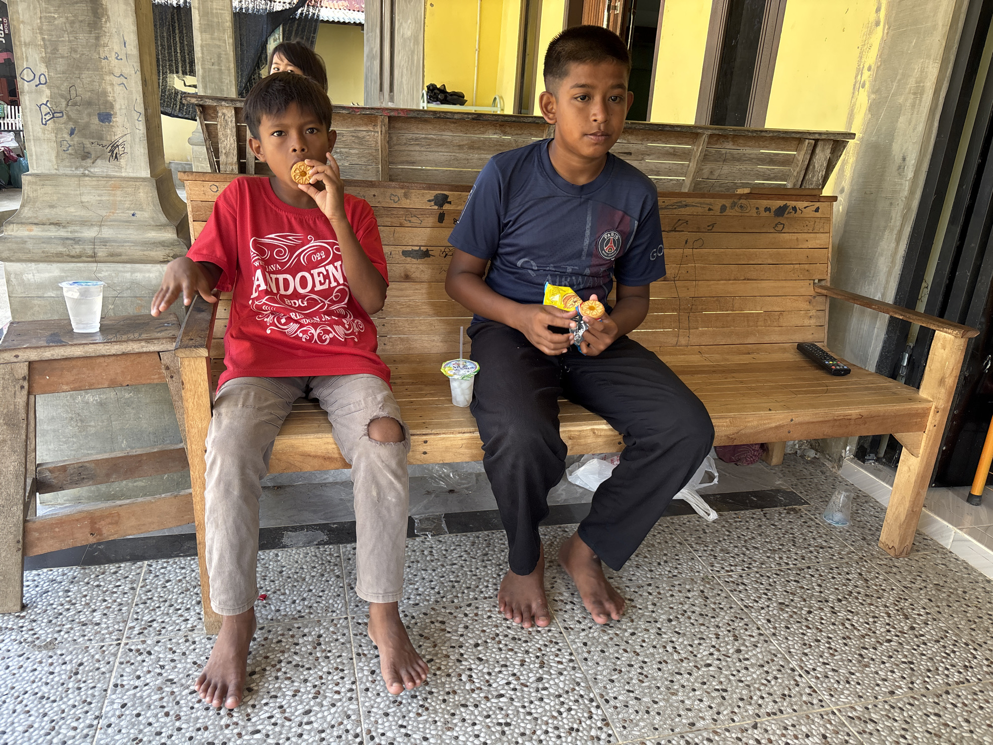 Two young boys are sitting on a wooden bench outdoors. The boy on the left, wearing a red t-shirt, is eating a cookie. The boy on the right, wearing a dark blue t-shirt, is holding a snack package and a cookie. They are barefoot, and the ground is tiled with small pebbles. In the background, there is a yellow building and part of a wooden structure. A remote control is visible on the bench. The image appears to have been taken in Lhokseumawe, Indonesia.