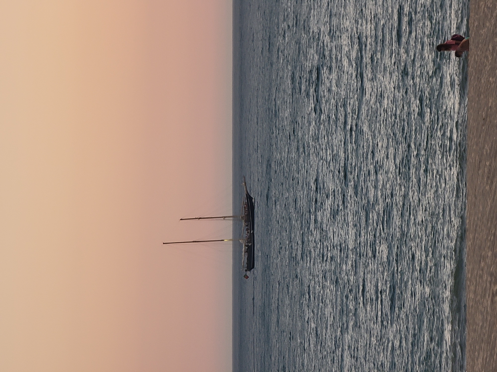 A photograph taken at dusk in Quarteira, Portugal, captures a serene maritime scene. A large, two-masted sailboat is positioned in the middle distance on a calm, deep blue ocean. The sky above is a soft gradient of peach and pink, indicative of twilight. In the foreground, on a sandy beach, a person wearing a pink top and a backpack is seated near the water's edge. The image conveys a sense of tranquility and the end of a day by the sea.