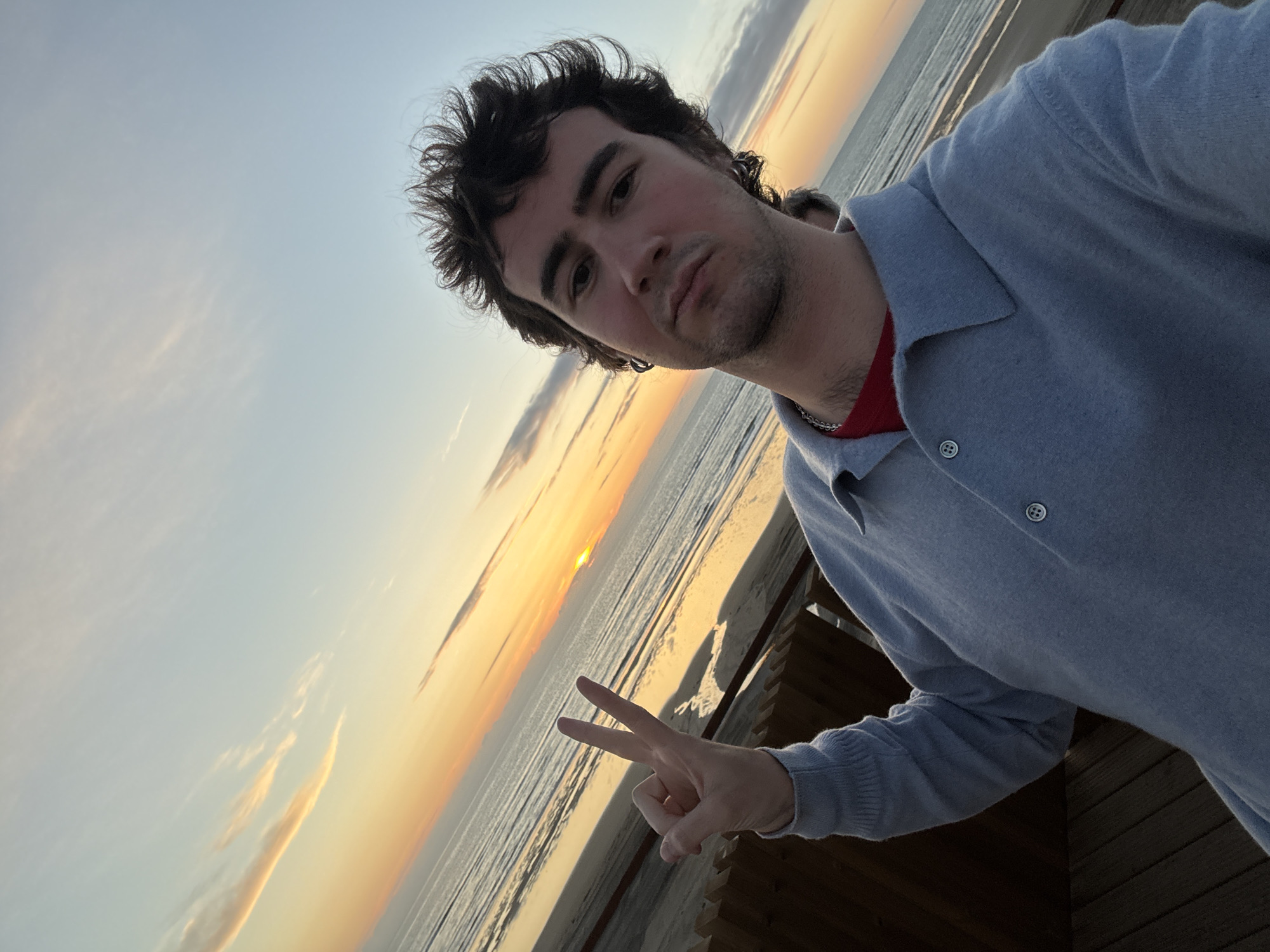 A young man is posing for a selfie at sunset on a beach in Soulac-sur-Mer, France. He has dark hair and is wearing a light blue sweater over a red shirt. He is making a peace sign with his right hand. The sun is setting over the ocean in the background, casting a warm orange glow on the scene. The wooden deck or boardwalk is visible in the foreground.