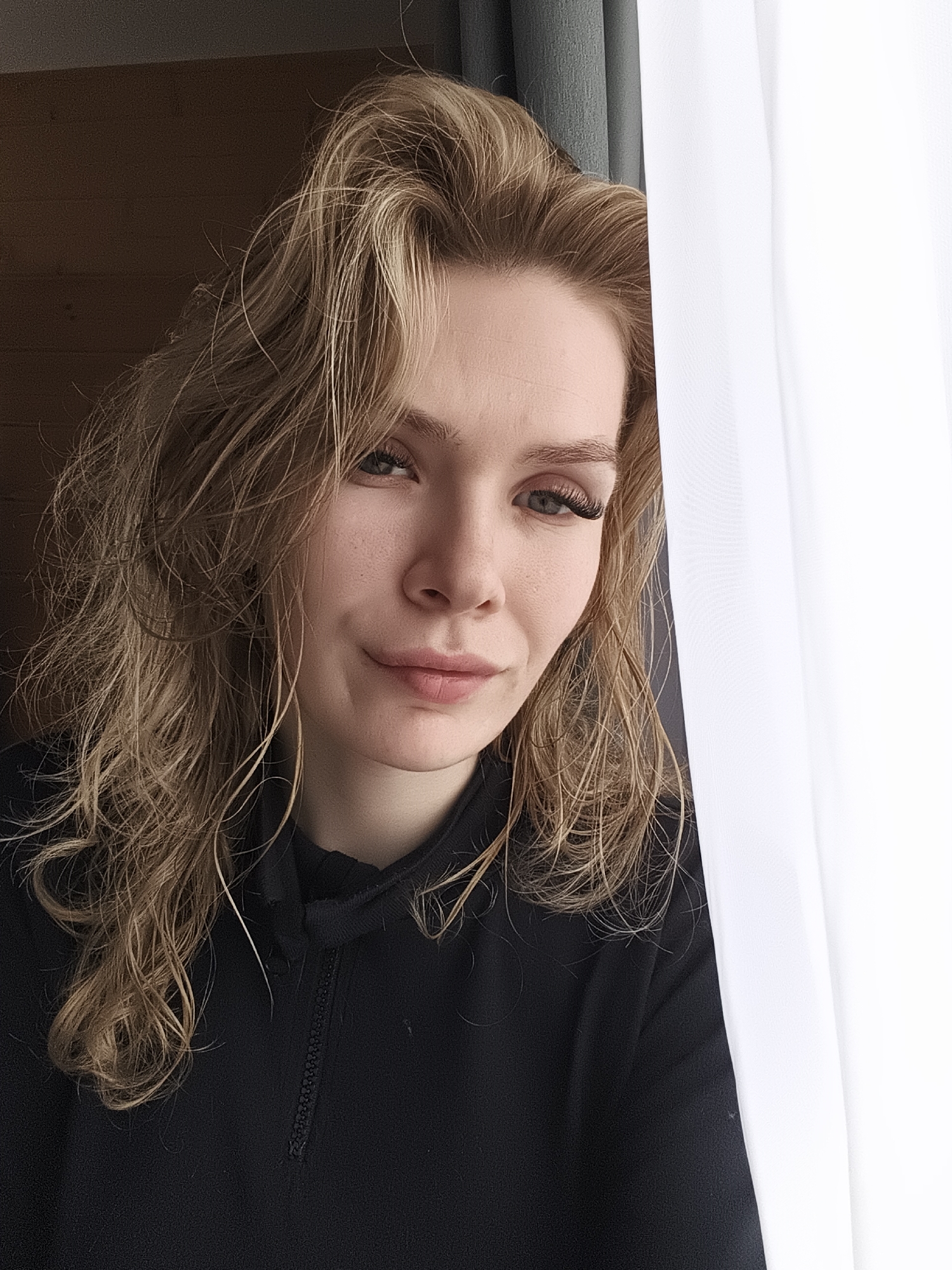 A close-up portrait features a young woman with long, wavy, light brown or blonde hair, appearing slightly unstyled or damp. She looks directly at the camera with a calm, confident expression, a subtle, almost smirking smile on her lips. Her eyes are light-colored, and she has prominent eyelashes. She is wearing a black top with a quarter-zip collar.
The image is framed on the right by a bright white, sheer curtain or fabric, which partially obscures the subject. To the left and behind her head, a darker, possibly gray or blue curtain is visible, contrasting with a warm-toned wooden panel or wall further to the left. The soft, natural lighting suggests a daytime setting, likely indoors near a window. The overall scene conveys a relaxed atmosphere. No text is visible in the media. The location context for this image is Русский Вожой, Russia.