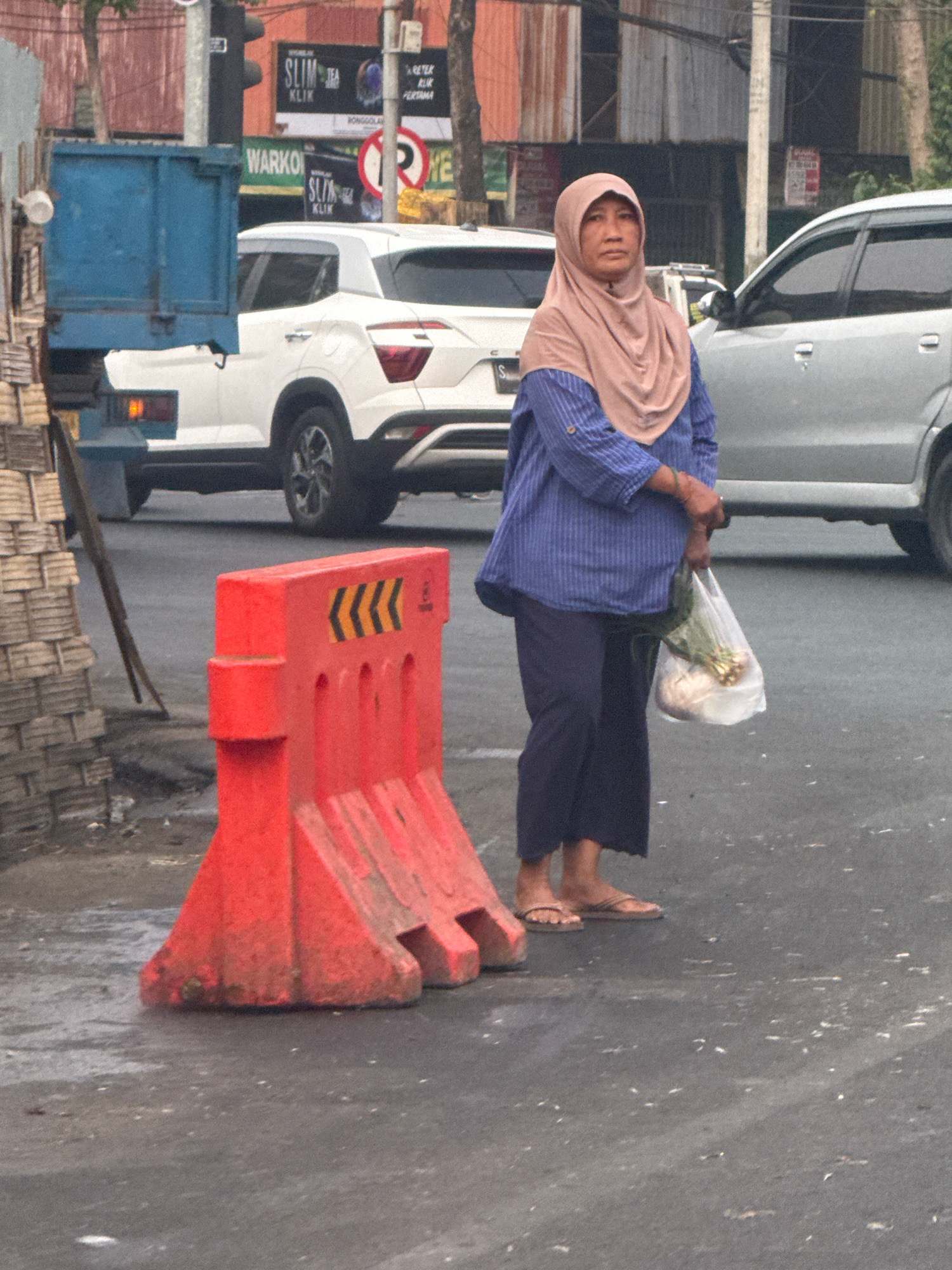 In Surabaya, Indonesia, a woman wearing a peach-colored hijab and a blue striped long-sleeved shirt stands on a paved road. She holds a translucent plastic bag filled with produce, possibly scallions and onions, in her right hand. Her dark blue pants are slightly frayed at the bottom. She wears brown flip-flops. In the foreground, a bright orange traffic barrier with black chevron markings is positioned on the road. Behind her, a white SUV and a light-colored car are visible. A blue truck, partially obscured by the barrier, is parked to the left. The background shows buildings with corrugated metal siding and trees. Traffic lights and signs are also present in the distance. The road surface appears damp.