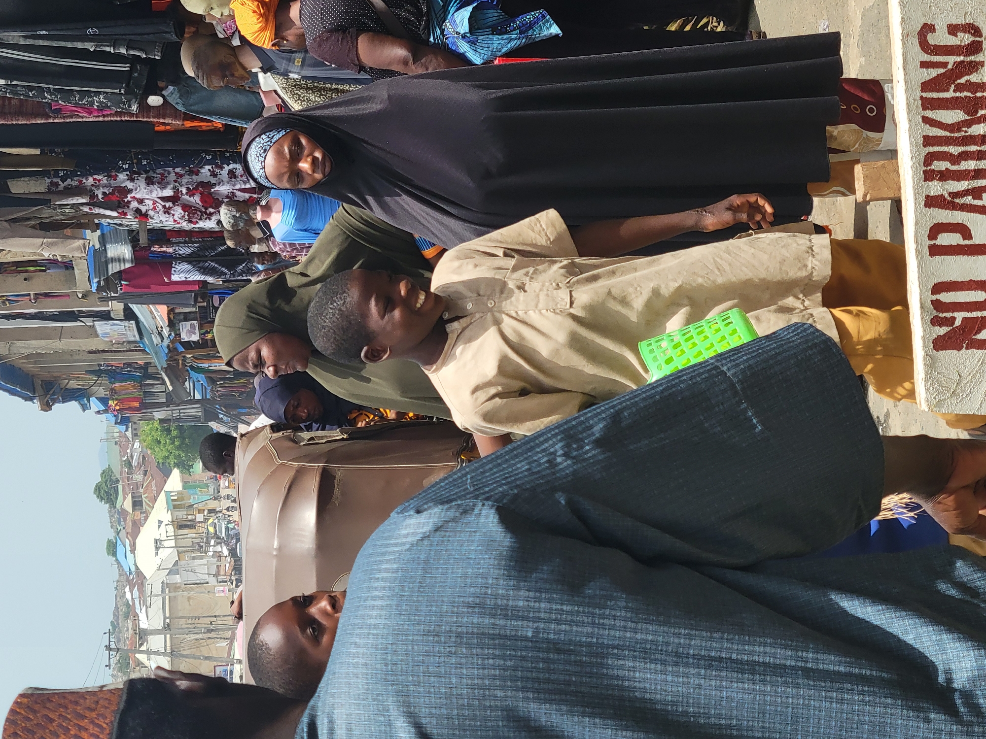In Suleja, Nigeria, a street scene features a vendor selling clothing and other wares. A young boy in a light brown shirt stands smiling, positioned between women in headscarves, with one woman wearing a black niqab. A man in a blue patterned shirt is present in the foreground. In the background, there is a street scene visible including buildings and a tricycle. A "NO PARKING" sign is partially visible on the right side.