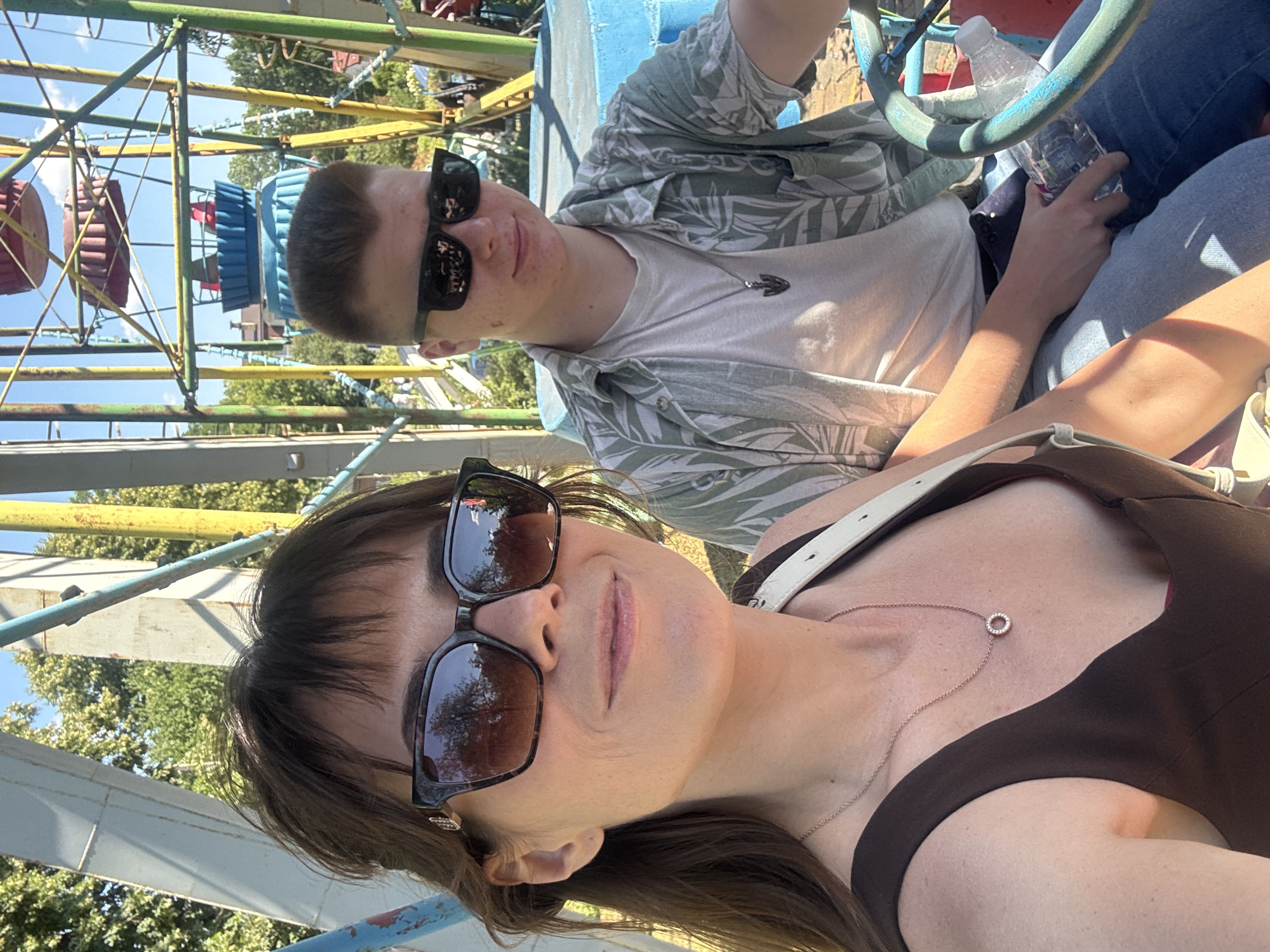 A woman and a young man are sitting in a ride at an amusement park. The woman is in the foreground, wearing a dark brown sleeveless top and a gold necklace with a circular pendant. She has brown hair and is wearing large, dark-rimmed sunglasses. The young man is behind and to her right, wearing a light-colored t-shirt and a green and white floral print short-sleeved button-down shirt, along with blue jeans and dark sunglasses. He has short, dark hair and is wearing a silver pendant necklace. They are both in a brightly colored open-air vehicle. The background shows the structure of the amusement park ride, with yellow and blue poles and red and blue circular seats. Trees and a clear blue sky are visible behind the ride. The overall scene suggests a sunny day at an amusement park.