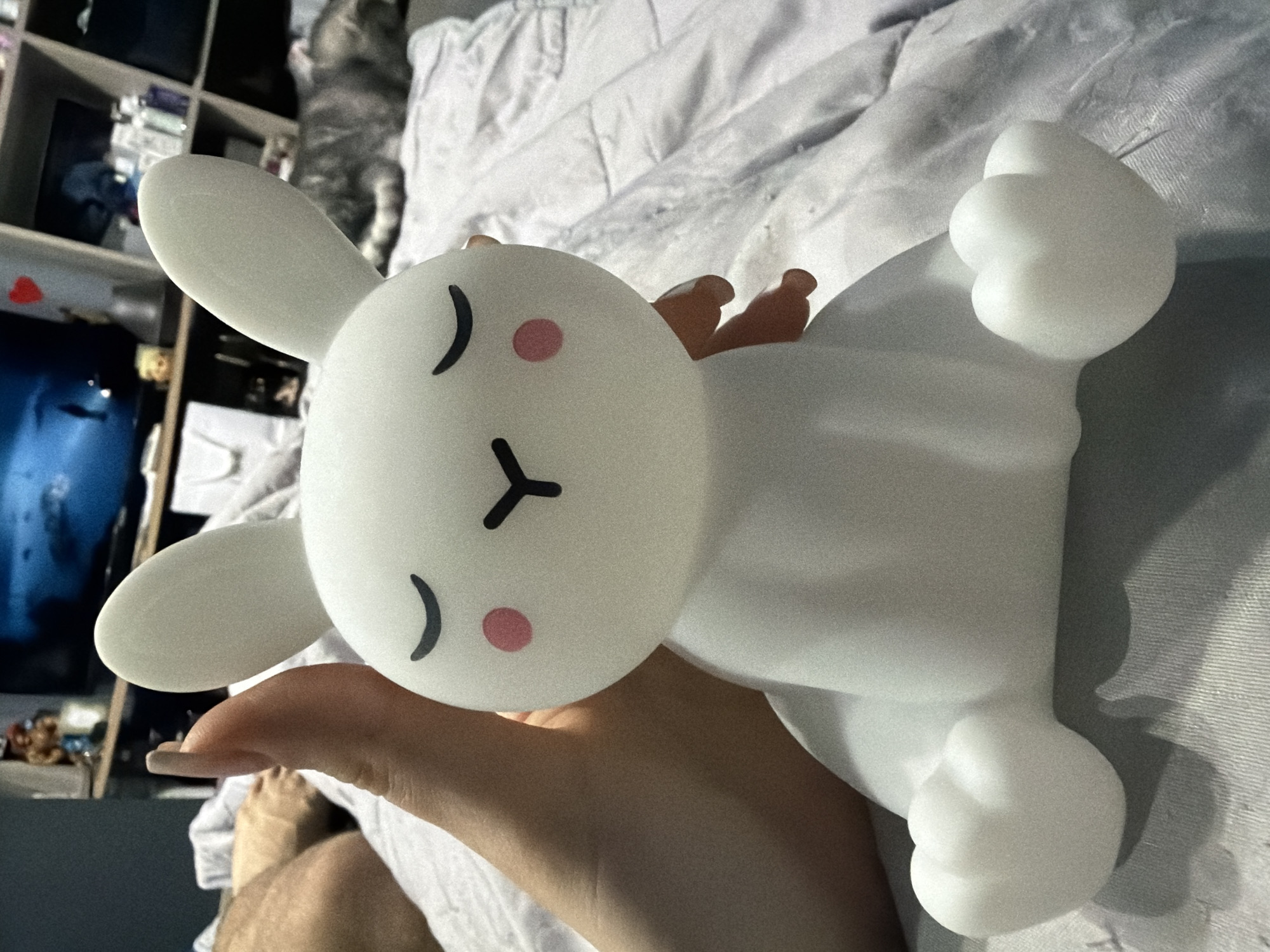 A close-up shot shows a person holding a white, cartoon-style rabbit-shaped night light in their hand. The rabbit has a simple, minimalist design with a round head featuring closed eyes (indicated by curved lines), pink cheek dots, and a small "Y" shape for the nose and mouth. The body of the rabbit is also white and bulbous, with visible arms and legs. The background is slightly blurred, revealing a bed covered with a white, textured blanket. There's a bookshelf visible to the left, containing various items. There is no information in the image to suggest a specific location in Burgas, Bulgaria.