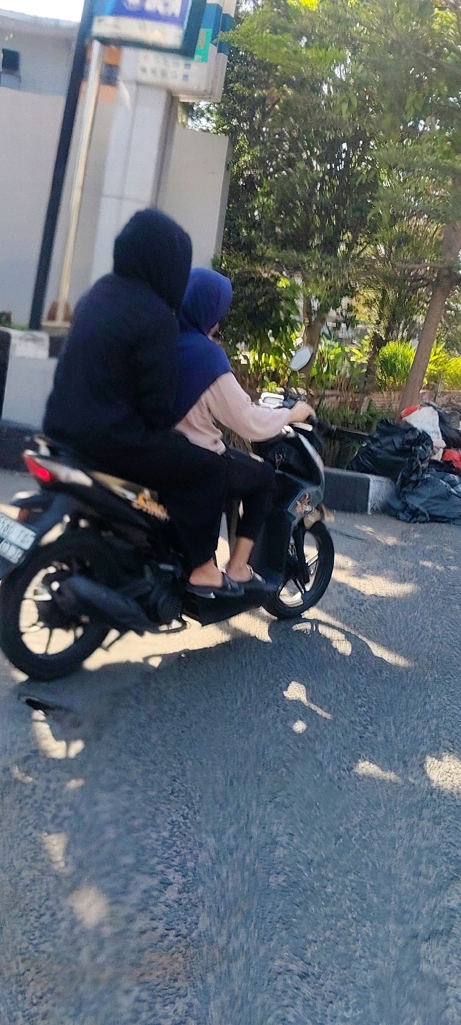 Two individuals, one operating a black scooter and the other as a passenger, are depicted on a street in Cibatu, Indonesia. The scooter is in motion, as indicated by the blurred background and the dynamic posture of the riders. The driver, in the front, is wearing a black hooded jacket, obscuring their face. The passenger, seated behind, is wearing a blue hijab and a light-colored top. The scooter has a license plate that appears to be B 5580 FAY, suggesting it is registered in Indonesia. The environment includes a street with a paved surface, a sidewalk, a pole with signage, and lush green trees. Adjacent to the sidewalk, there are several black trash bags. The lighting suggests it is daytime, with sunlight casting shadows on the road, indicating a clear or partly cloudy sky. The scene captures a common mode of transportation in Indonesian cities.