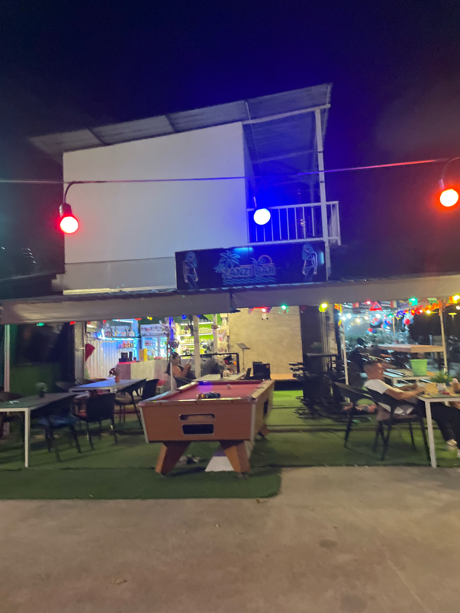 An outdoor bar scene at night in Pattaya City, Thailand, features a pool table in the foreground, an open-air bar with patrons seated at tables, and a second story with a balcony. Red and blue lights illuminate the scene, and string lights with flags are hung across the bar area. A man is seated at a table on the right side of the image, looking towards the right. The bar is decorated with images of palm trees and women. The ground is covered with artificial grass, and concrete pavement is visible in the foreground.