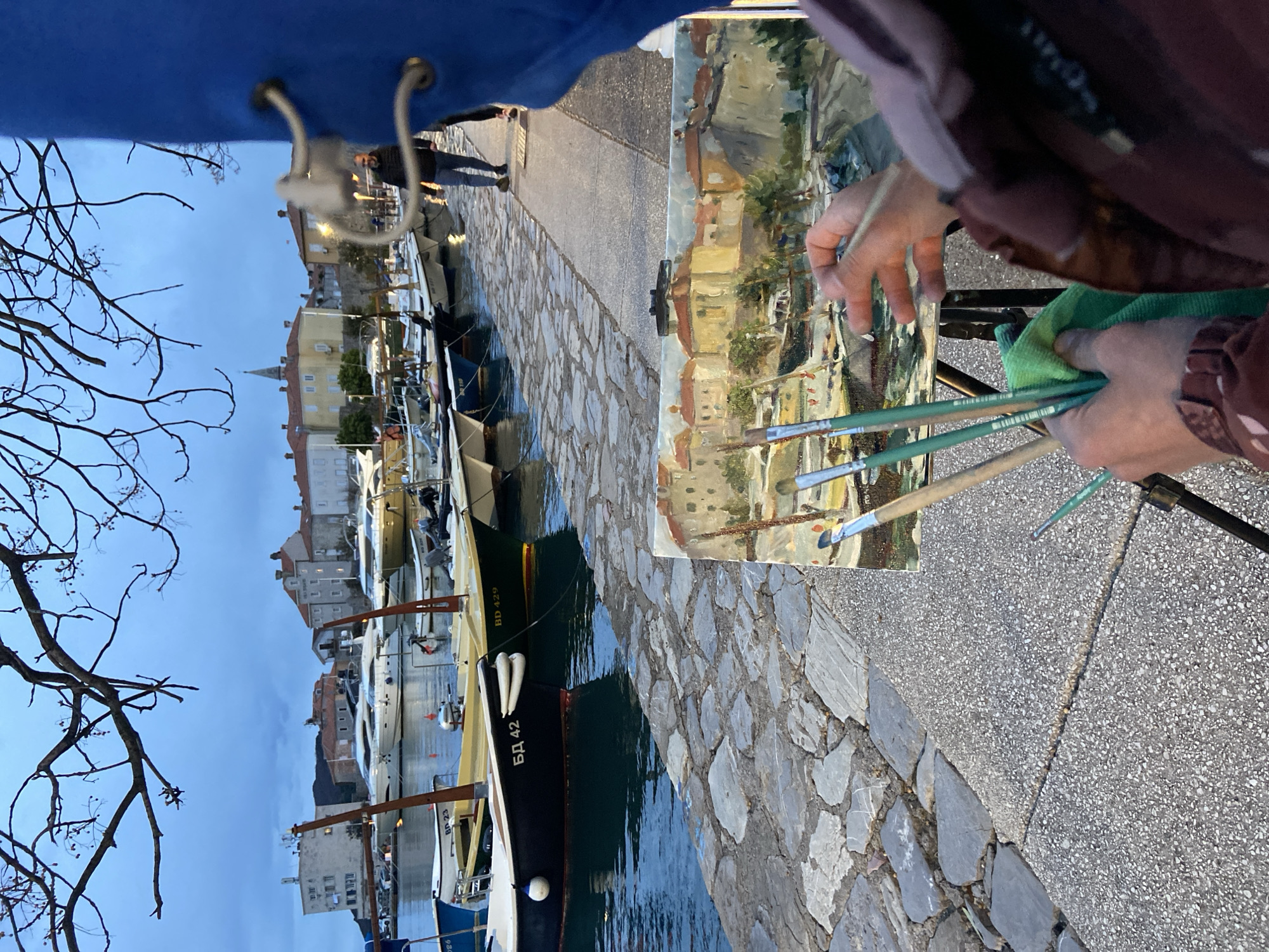 The image shows a person painting a plein air scene of the Budva harbor in Montenegro. The foreground features a cobblestone quay lined with several docked boats. The boats vary in size and color, some bearing Cyrillic script. In the background, a cityscape with buildings of varying heights and architectural styles can be seen. A bare tree branch is visible in the top-left corner. An individual stands on the quay in the distance. The painter is holding paintbrushes and a green cloth while working on a canvas mounted on an easel, capturing the harbor scene.