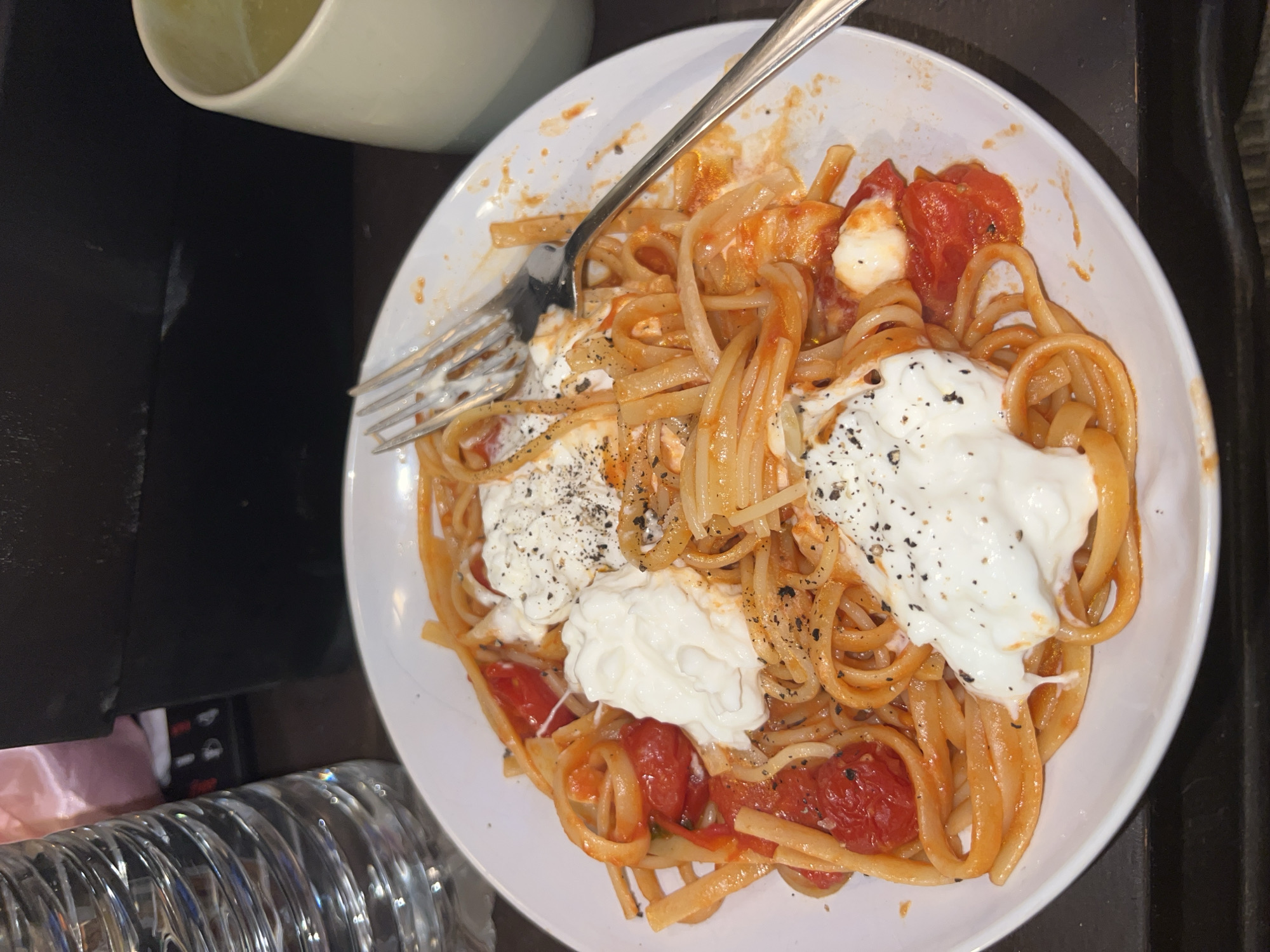 A plate of linguine pasta is presented on a white plate with a fork placed on top of the pasta. The pasta is topped with a red tomato sauce and dollops of white cream. Black pepper is visible on the cream dollops. To the left of the plate, there is a white mug and a clear water bottle. The scene is located in an indoor environment, possibly a restaurant or home setting. No evidence suggests a specific location in Paris, France.