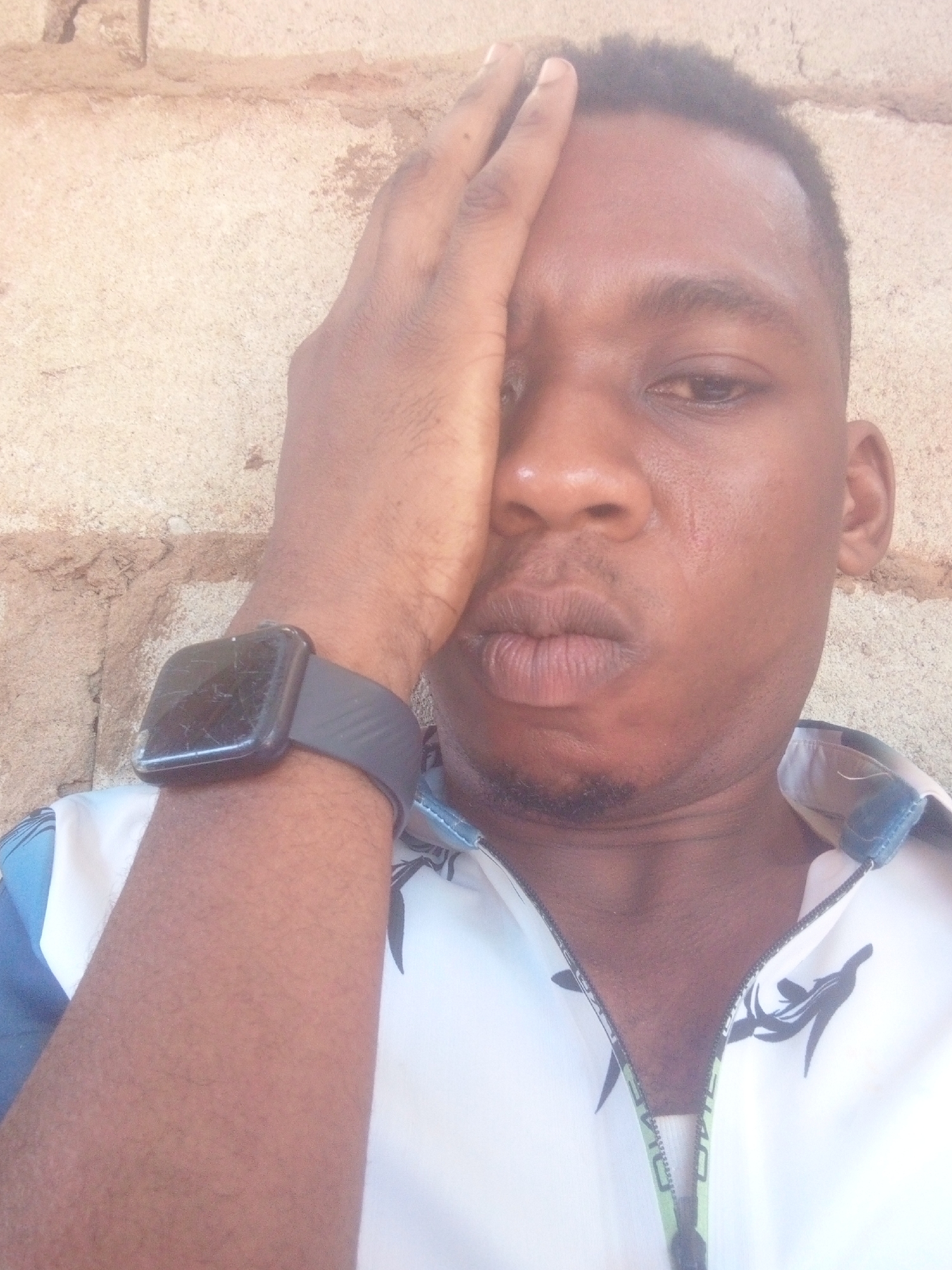 A man with brown skin poses for a close-up photo. He has dark, short hair and a neatly trimmed beard. His expression is somewhat neutral with slightly pursed lips. He has a black smartwatch on his left wrist. His left hand is raised, partially covering his face. He is wearing a light-colored shirt with a pattern. The background appears to be a textured wall, possibly made of concrete or stone. The location is unknown.