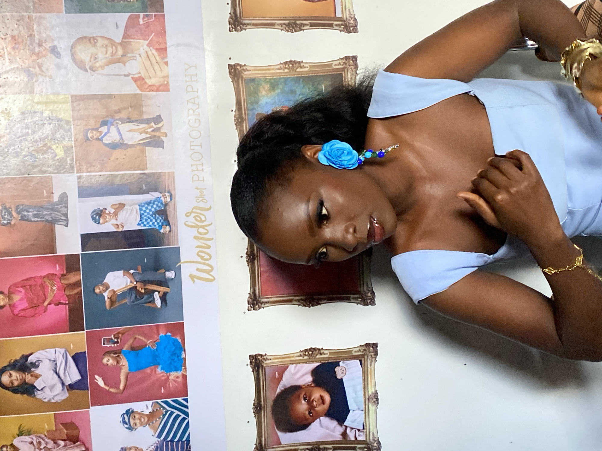 A young Nigerian woman is posing in front of a wall decorated with framed photos and a large banner. The banner displays the text "Wonder Shot PHOTOGRAPHY" in a decorative font. The framed photos appear to be portraits and studio shots, some featuring children in various outfits and poses. The woman is wearing a light blue dress and a large, vibrant blue flower earring with dangling beads. She is also wearing a gold bracelet. Her hair is pulled back into a ponytail. The background wall is white. The lighting suggests an indoor setting, likely a photography studio or a home decorated with studio backdrops. The provided context about the location in "Gwong, Nigeria" indicates the potential cultural and geographical setting of the scene.