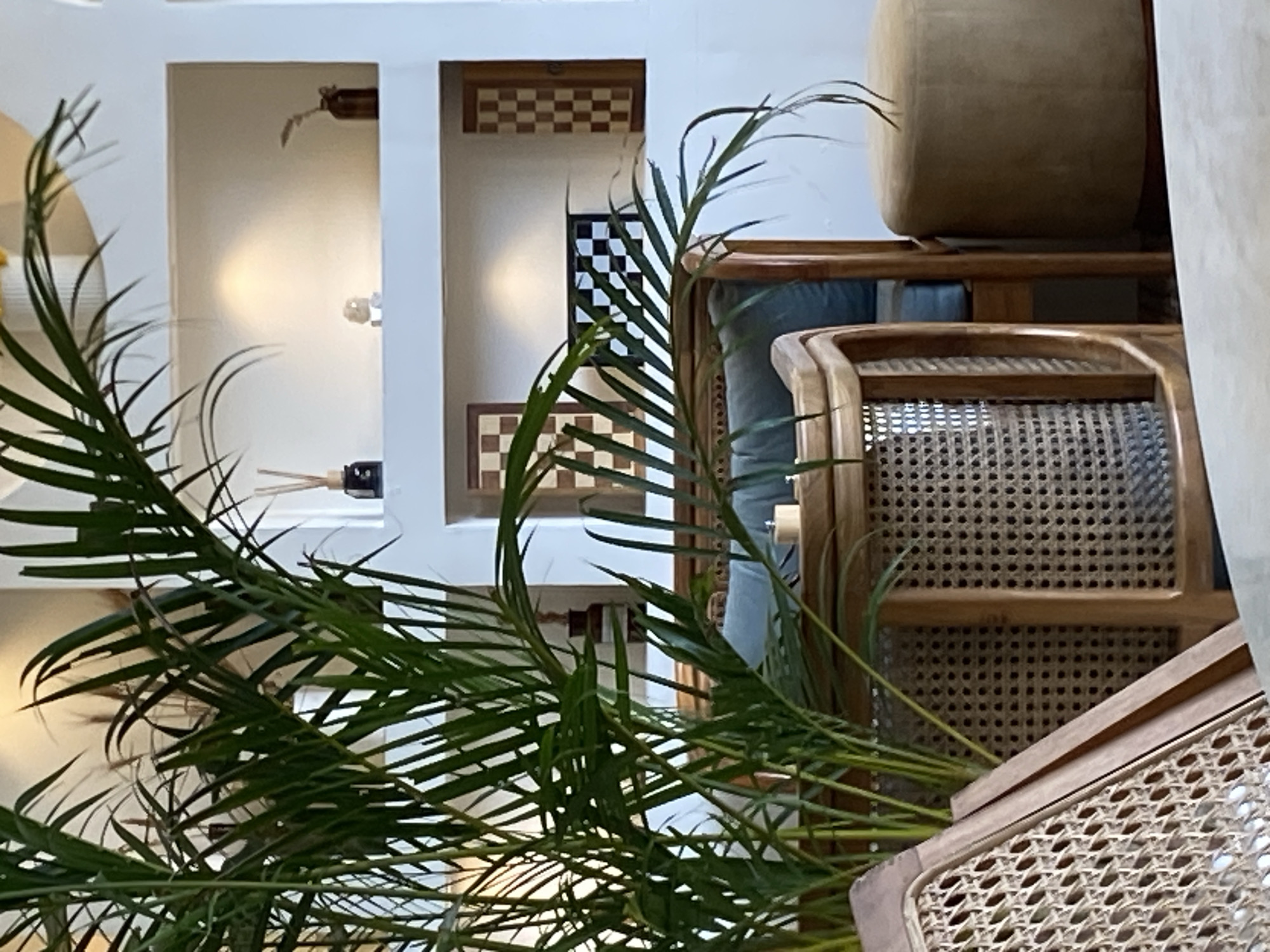 An indoor scene in North Kuta, Indonesia, shows a seating area with a large potted plant in the foreground. The plant's fronds are lush and green, partially obscuring a woven rattan chair with a blue cushion. Behind the chair is a light-colored wall with recessed shelves. These shelves display decorative items, including a diffuser with reeds, a small glass object, a vase with dried foliage, and several framed checkerboard patterns. A round, light brown ottoman is visible to the right of the rattan chair. The overall ambiance suggests a modern, minimalist interior design.