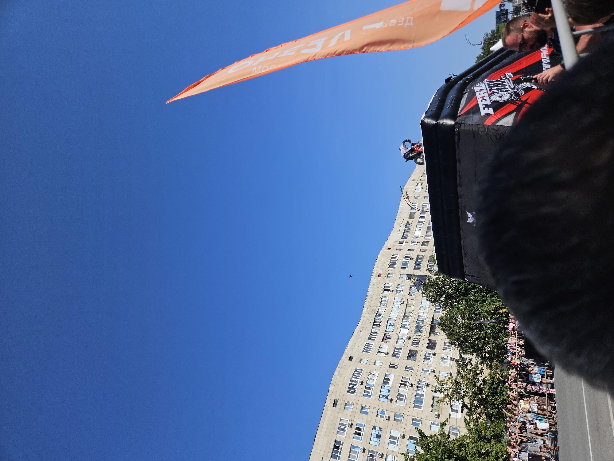 On a bright, clear day in Ulyanovsk, Russia, a motocross stunt rider prepares for a jump atop an inflatable ramp. The rider, dressed in a white and black checkered shirt, is positioned on a red dirt bike at the apex of the ramp. The ramp itself is black with red and white graphics, featuring the text "FERZ" and what appears to be a stylized wheel.
To the left of the ramp, a tall, multi-story apartment building with numerous windows and air conditioning units dominates the background. Lush green trees are visible at the base of the building and in the mid-ground.
In the foreground, a crowd of spectators stands behind a metal barrier, observing the event. Many are holding up their phones to capture the moment. An orange flag with black lettering, partially obscured by the ramp, stands tall to the right of the frame. A man wearing glasses and a black t-shirt is visible in the lower right corner, facing towards the ramp. The sky is a vibrant, cloudless blue.