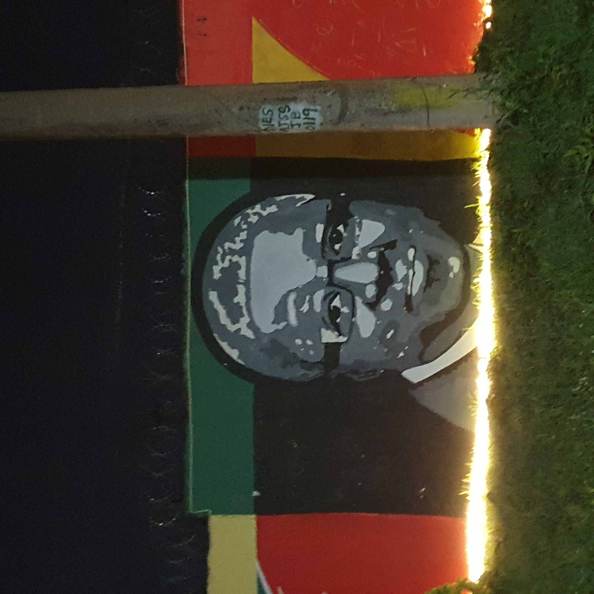 The image shows a mural of a man's face on a wall, likely in Barakin Lerawi, Nigeria. The mural is rendered in shades of gray and white, with the subject wearing glasses and having short, wavy hair. The background of the mural features horizontal bands of color: yellow, green, and black. To the right of the mural, a concrete pole is visible, with white graffiti on it that appears to read "NES AJSS JB 0119". The wall is bordered at the top by barbed wire. In front of the wall, there is a strip of bright, yellowish light, possibly from streetlights, illuminating the grass and the lower part of the mural. The overall scene is dimly lit, suggesting it was taken at night.