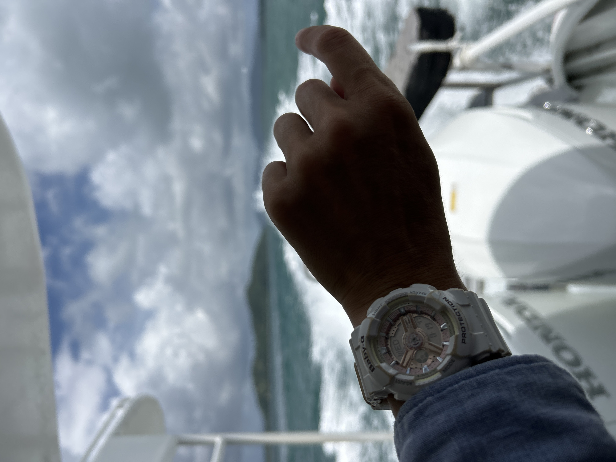 A person's hand wearing a white Baby-G watch is held up against a blurred background of the ocean and islands. The watch has a rose gold face with the day of the week and time displayed. The person is wearing a blue long-sleeved shirt. The background shows choppy water, a boat, and distant islands under a cloudy sky. The scene appears to be a boat ride in Ratsada, Thailand.