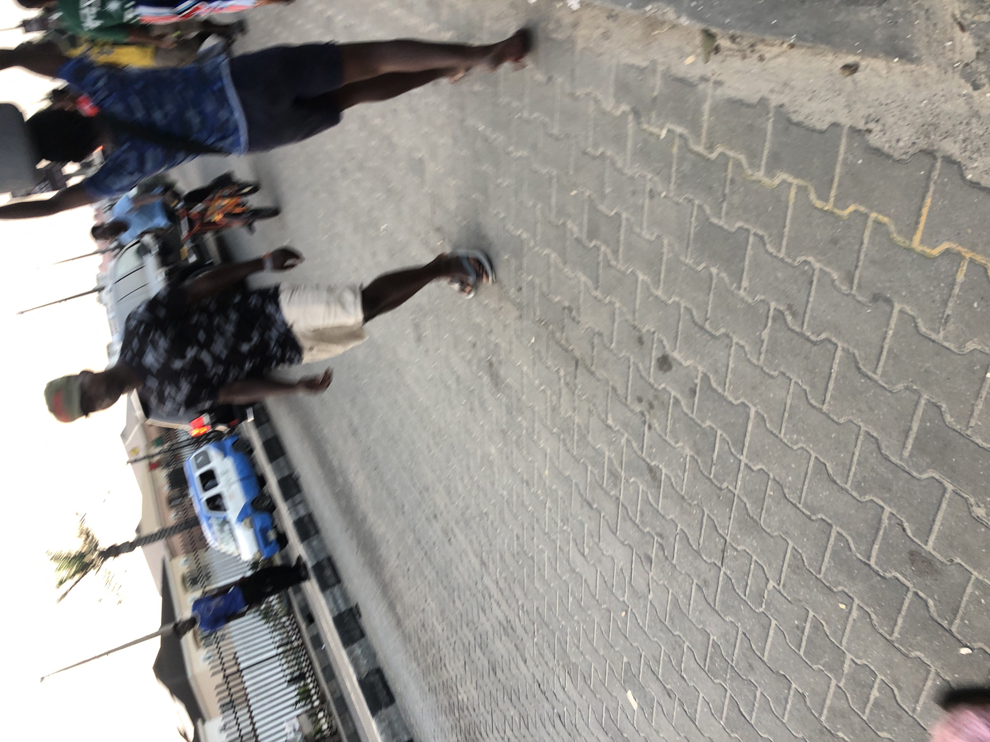 The image depicts a street scene in Moba, Nigeria. The ground is paved with interlocking gray bricks. Two individuals are visible, seemingly walking on the pavement. One is wearing a dark patterned shirt and light-colored shorts, while the other is wearing a blue patterned shirt and dark shorts. A blue and white vehicle, possibly a bus, is parked on the side of the street. There are trees in the background, and buildings are visible in the distance. A metal fence separates the pavement from the street.