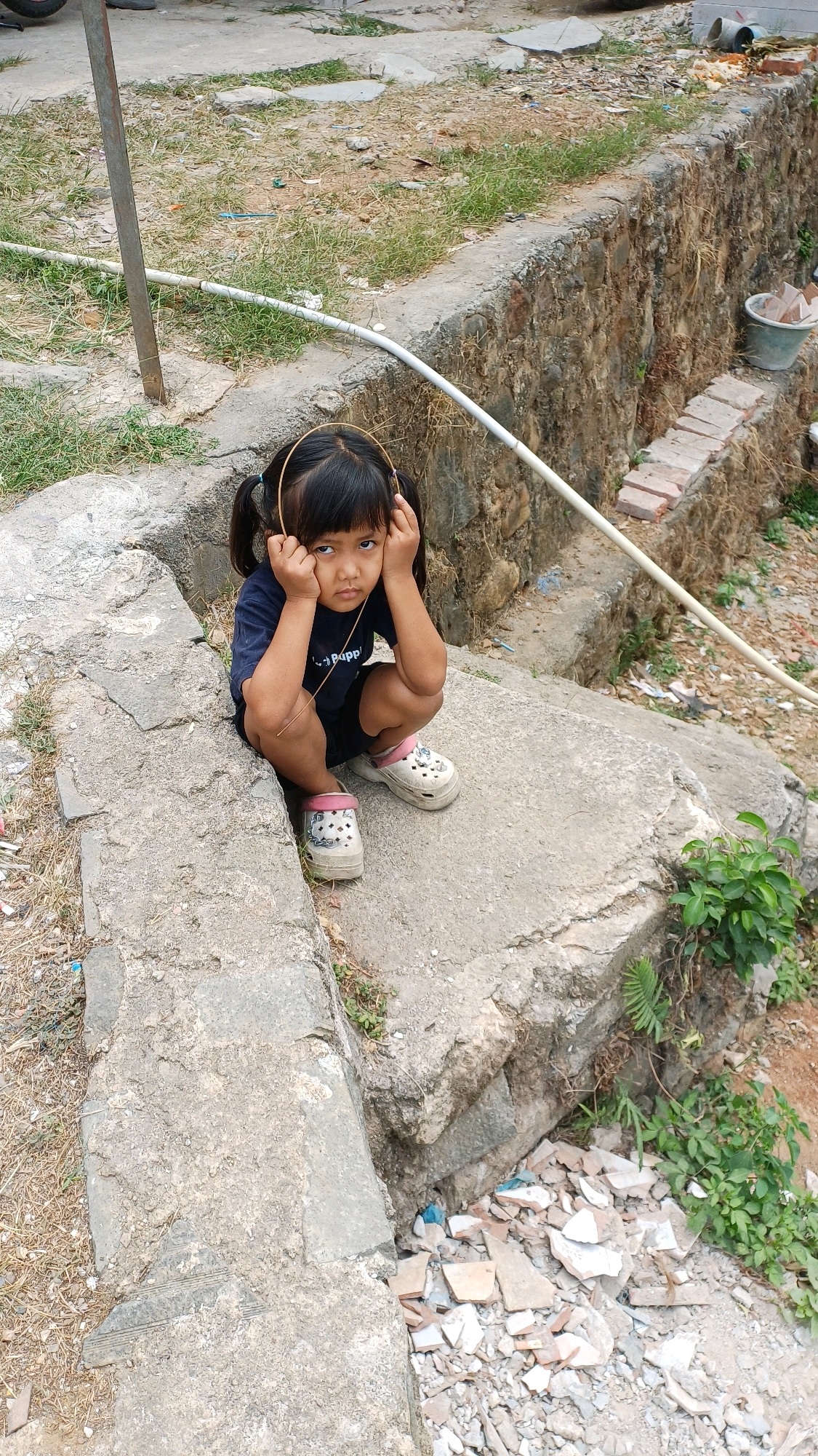 A young girl with black hair tied in pigtails is squatting on concrete steps, her hands covering her ears. She is wearing a dark blue t-shirt with white text and dark shorts. Her shoes are white with pink accents. The surrounding environment appears to be an outdoor area with rough concrete, dirt, and sparse vegetation. Debris, including broken tiles, is scattered on the ground. A white pipe runs along the edge of the concrete structure. The scene suggests an undeveloped or construction area. The photograph was taken in Gunung Putri, Indonesia.