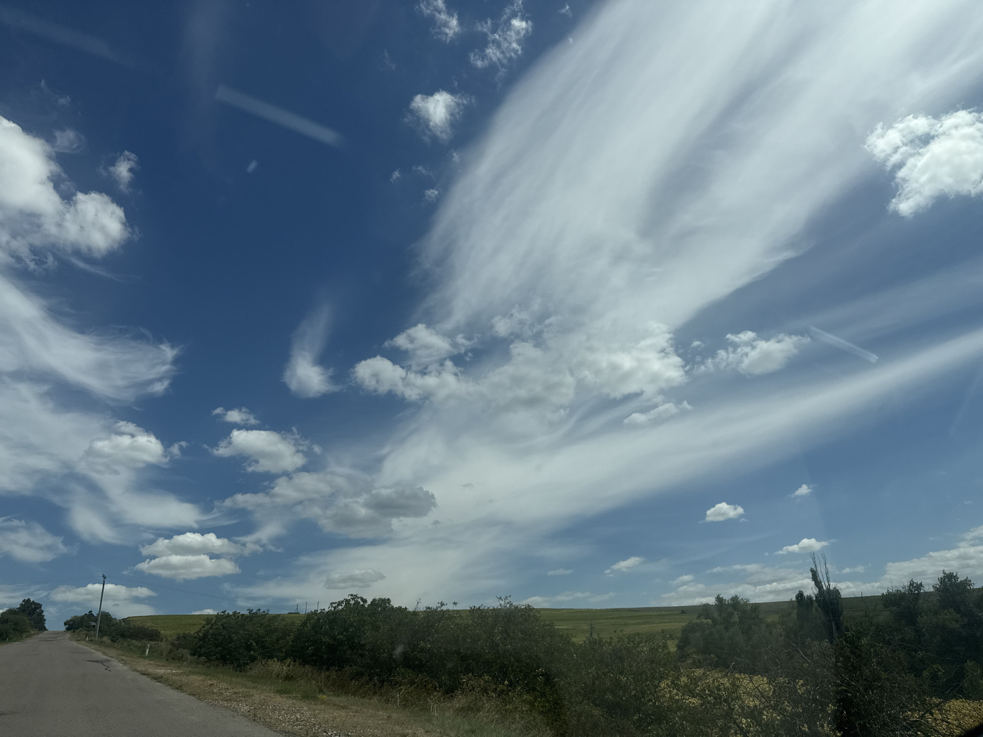 The image depicts a rural landscape under a dynamic sky. A paved road curves from the lower left towards the middle ground, bordered by dry grass and sparse vegetation. On the right side of the road, a line of dense green bushes and trees forms a natural barrier. Beyond this, rolling hills covered in green fields stretch towards the horizon. The sky is a vibrant blue, punctuated by a dramatic display of clouds. Wispy, elongated clouds stretch across the upper right portion of the frame, appearing as if painted strokes. Scattered lower clouds, some fluffy and white, are present throughout the sky. In the far distance, the faint silhouette of a utility pole is visible on the left side of the road, and another less distinct structure or tree is seen on the far right. The overall scene conveys a sense of open space and natural beauty, possibly captured during daylight hours with good weather conditions. There are no people or animals visible in the frame, and no specific man-made structures other than the road and utility pole are prominent.