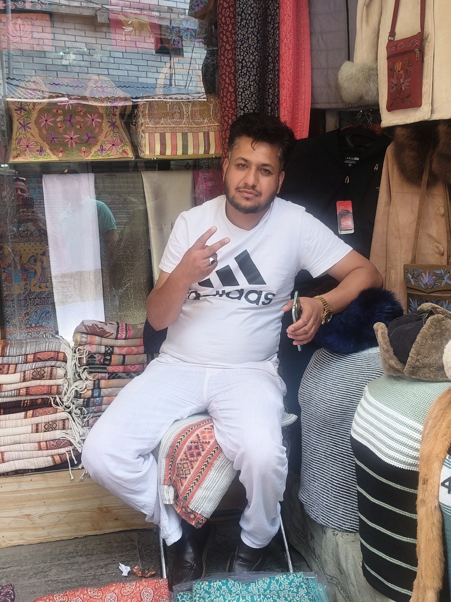A man in white clothing sits in front of a store display in Vashisht, India. He wears a white t-shirt with an Adidas logo, white pants, and black shoes. He holds a smartphone in his right hand and makes a peace sign with his left hand. The store display behind him features stacks of textiles, including colorful woven blankets and embroidered fabrics. Garments like jackets and purses are also visible, along with patterned curtains and a brick wall. The overall impression is that of a street-side vendor's stall.