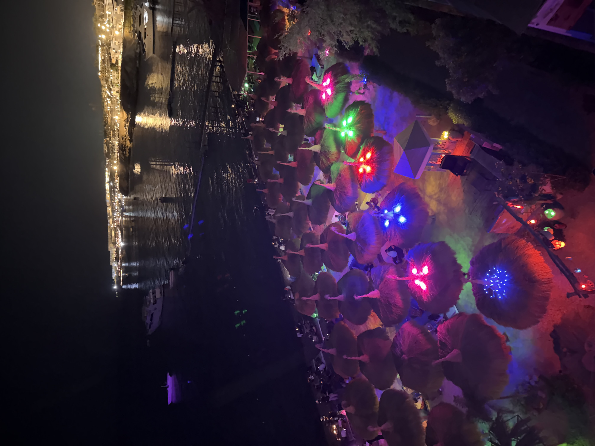This is a nighttime aerial view of a beach in Sharm El Sheikh, Egypt, likely along Naama Bay. The scene depicts a beach area illuminated by colorful lights. Dozens of thatched umbrellas are arranged on the sand, many adorned with vibrant LED lights in red, green, blue, and pink. Some of these umbrellas are illuminated from underneath, casting a glow on the sand and surrounding areas.
In the background, the dark water of the Red Sea is visible, with reflections of lights from buildings and boats. Several boats and yachts are anchored in the bay, and a distant shoreline is lined with illuminated buildings and structures, characteristic of a resort town. Small groups of people are visible scattered on the beach, some seated under the umbrellas, suggesting a lively evening atmosphere.
There is a small gazebo-like structure with a table and chairs in the foreground, also illuminated. The overall impression is that of a well-lit, festive beachside venue at night.