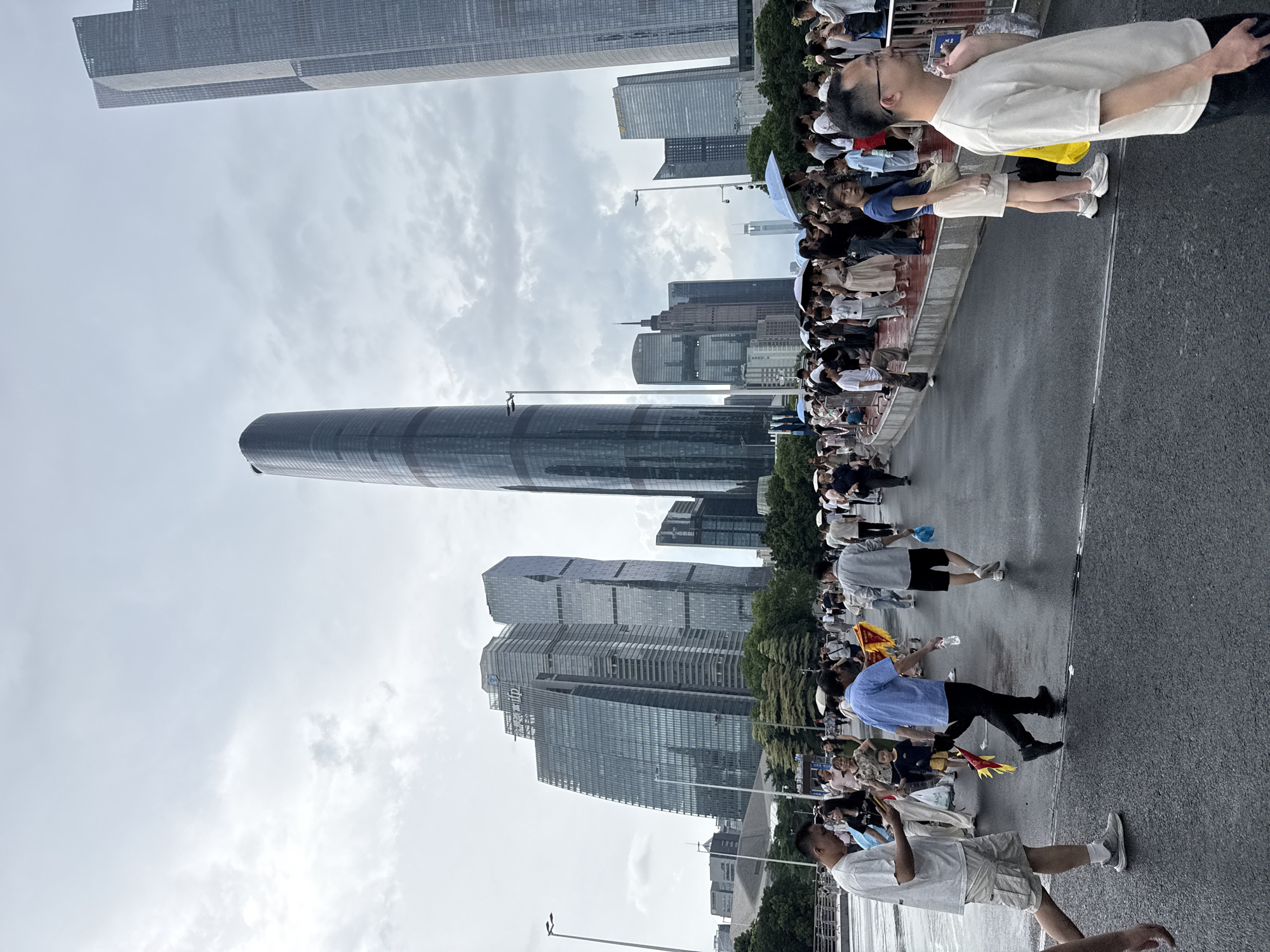 This image captures a street scene in Tianhe District, China, on a cloudy day. A large crowd of people is gathered on a wet asphalt surface, which appears to be a pedestrian walkway or street. In the background, a cluster of modern skyscrapers dominates the skyline. The architecture of these buildings is characterized by glass facades and varied geometric shapes. One particularly tall, cylindrical building stands out, while others are more rectangular.
The people in the foreground and midground are walking and standing, some holding umbrellas. Their clothing suggests a casual setting, with many wearing t-shirts and shorts. A few individuals are holding small, colorful flags or banners. The ground is wet, indicating recent rain or a damp environment. The overall atmosphere is one of public gathering, possibly for an event or simply navigating the urban space. The sky is overcast with diffused light, contributing to a muted color palette.