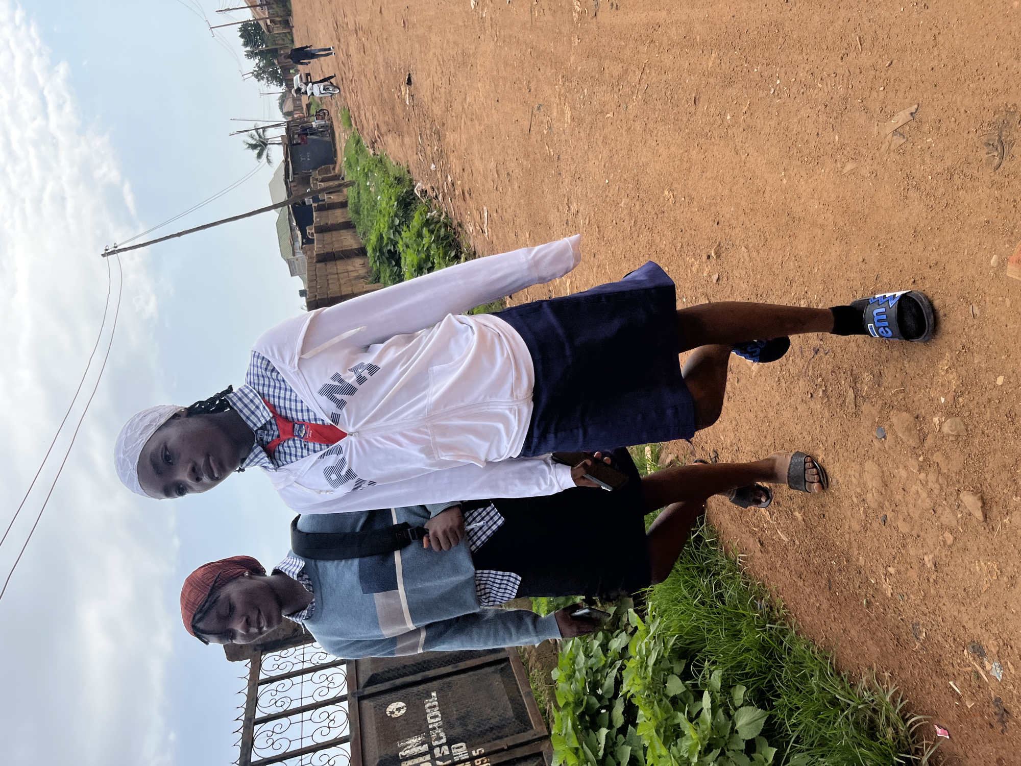 Two young girls, appearing to be students in uniform, walk along a dirt road in an urban environment, likely in Jos, Nigeria. The girl in the foreground wears a white zip-up hoodie over a blue and white checkered shirt and a dark blue skirt, with black slides on her feet. She has a white headscarf and is looking to her left. The girl behind her wears a blue and grey striped sweater over a blue and white checkered shirt and a black skirt, with a red headscarf. She carries a backpack over her left shoulder and is also looking to her left. They are walking away from the viewer. The road is unpaved and dusty, with a low brick wall and green vegetation along the left side. In the background, there are several buildings, utility poles with wires, and other people and vehicles, including a motorcycle. The sky is cloudy. The scene suggests an ordinary day in a residential or semi-commercial area of the city.