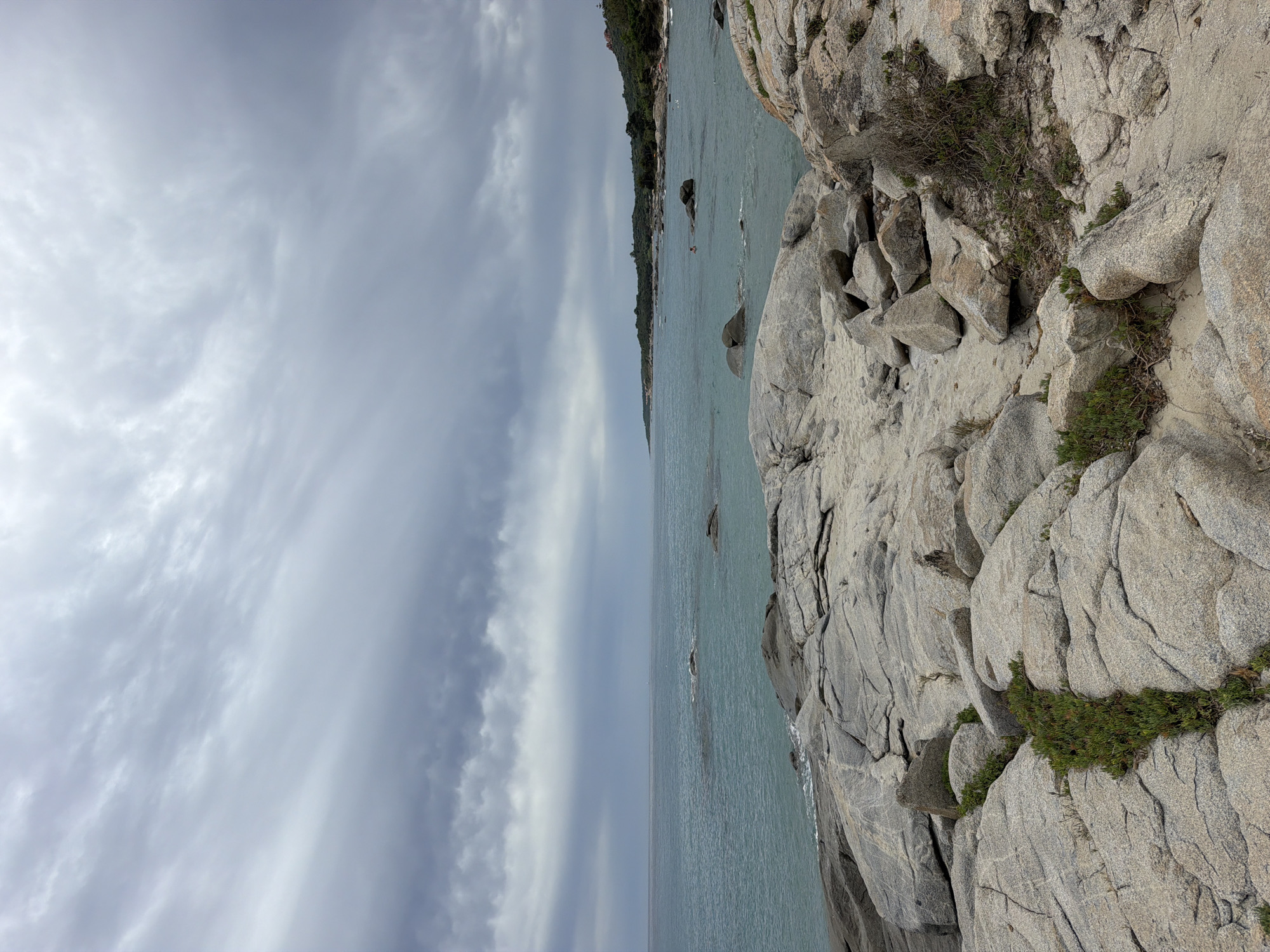The image depicts a rocky coastline with a calm, turquoise sea under a cloudy sky. In the foreground, large, weathered granite rocks dominate the scene, with sparse green vegetation growing in crevices. The sea stretches to the horizon, where it meets a distant coastline dotted with trees and hills. The sky is filled with heavy, grey clouds, suggesting overcast weather. A person is visible swimming in the sea in the middle ground. This is likely the coast near Porto Frailis in Tortolì, Italy, a well-known area for its rocky shores and clear waters.