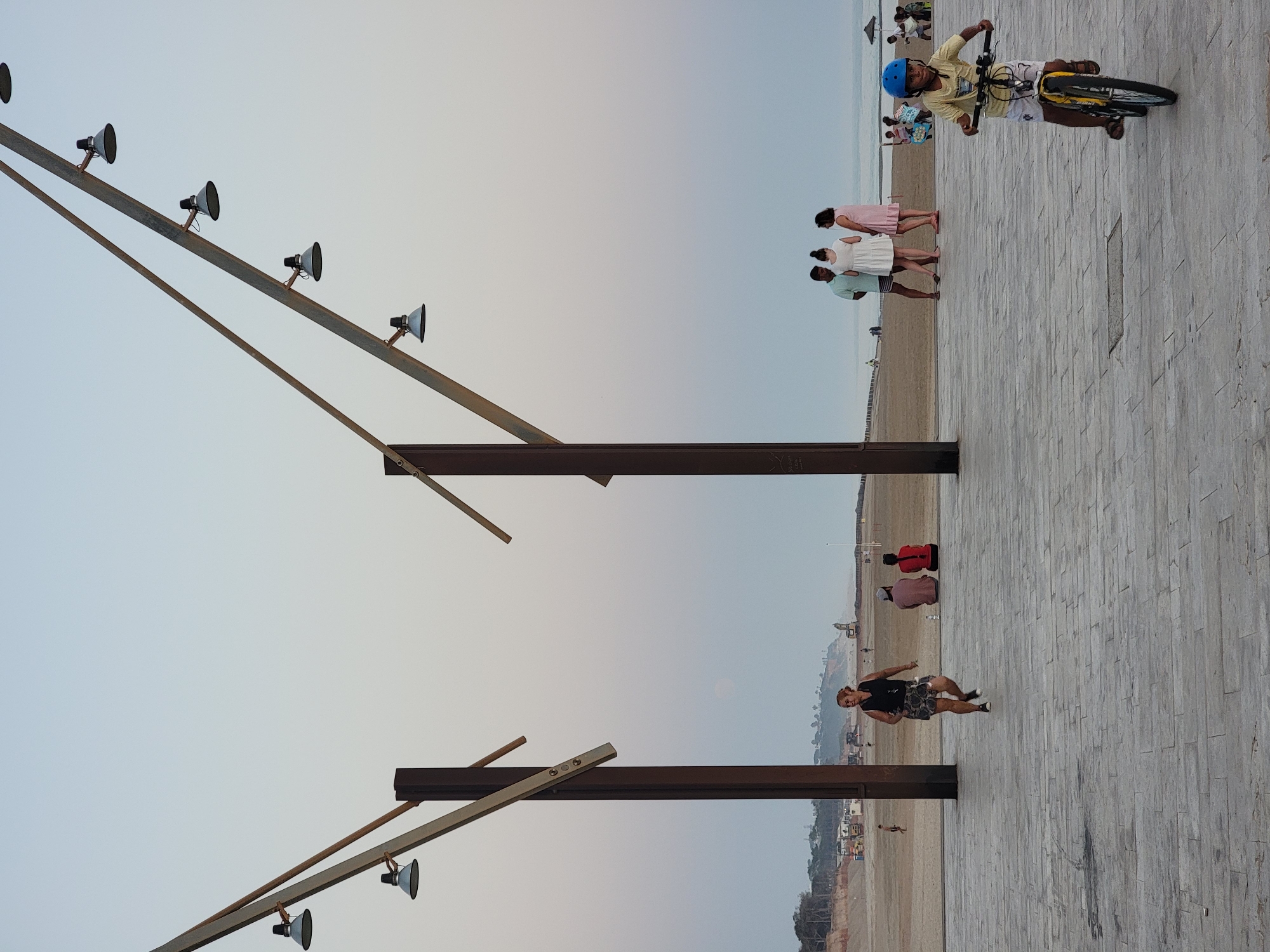 An eye-level, medium shot captures a wide, paved promenade with a sandy beach and the Atlantic Ocean in the background, at dusk in Quarteira, Portugal. Two large, metal, angled structures with multiple light fixtures extend into the sky from the left and right sides of the frame, framing the scene. In the foreground, a young boy wearing a blue helmet and a yellow t-shirt rides a yellow bicycle towards the right. His body is angled slightly to his left, and his head is turned towards the viewer. Further back, on the promenade, a woman with blonde hair tied in a bun, wearing a black tank top and patterned shorts, runs towards the left. Her head is turned towards her right. A group of three people, two women and a man, stand on the promenade facing away from the camera, conversing. The man wears a mint green shirt and striped shorts, one woman a white dress, and the other a pink dress. Behind them, the sandy beach is populated with several individuals, some sitting and some walking. In the middle ground, the horizon shows a coastline with buildings and vegetation. The sky is a pale, gradient blue. The pavement is made of large, rectangular grey tiles.
