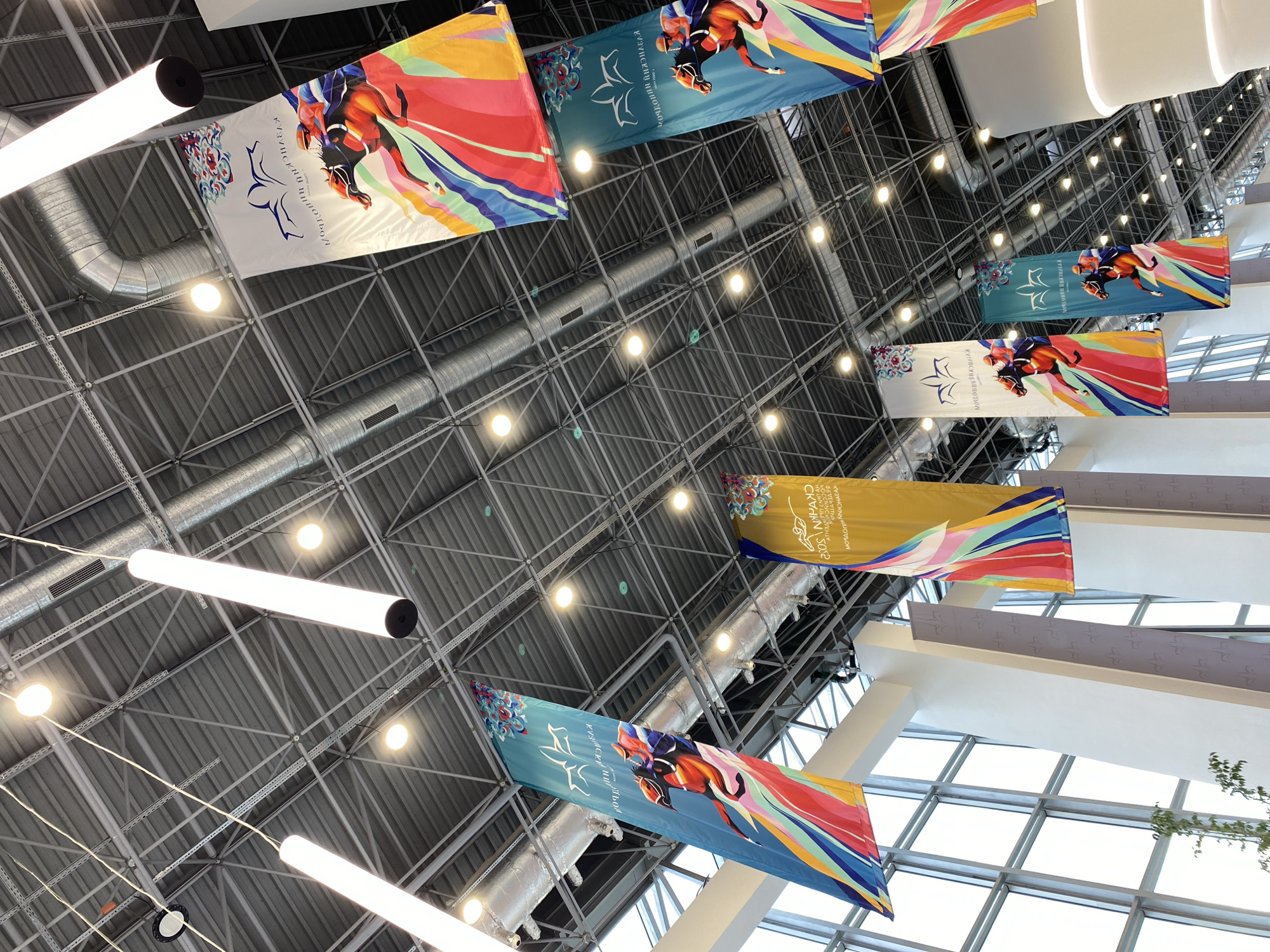 This media file is a photograph taken from below, looking up at the interior of a large, modern building with a high, exposed metal ceiling structure. Long, cylindrical white lights are suspended from the ceiling, along with large banners that are also hung from above. The banners feature vibrant, colorful abstract designs and illustrations of horses in motion, with some displaying text.
The scene appears to be an indoor exhibition space, convention center, or arena, judging by the scale of the structure and the prominent banners. The banners suggest an event related to horses or equestrian activities. Some of the banners have text that appears to be in Cyrillic script, with one possibly reading "КАЗАНСКИЙ ИПОДРОМ" which translates to "Kazan Hippodrome," indicating the location is likely in Kazan, Russia. Another banner appears to have text that includes "SKHYN 2022," suggesting a specific event or competition.
The lighting is bright, likely from the artificial lights and possibly natural light filtering in from windows or skylights not fully visible in the frame. The overall impression is one of a well-lit, spacious, and modern venue hosting a significant event. There are no people directly visible in the image, and the focus is on the architectural elements and the decorative banners. The time of day and weather are not discernible.