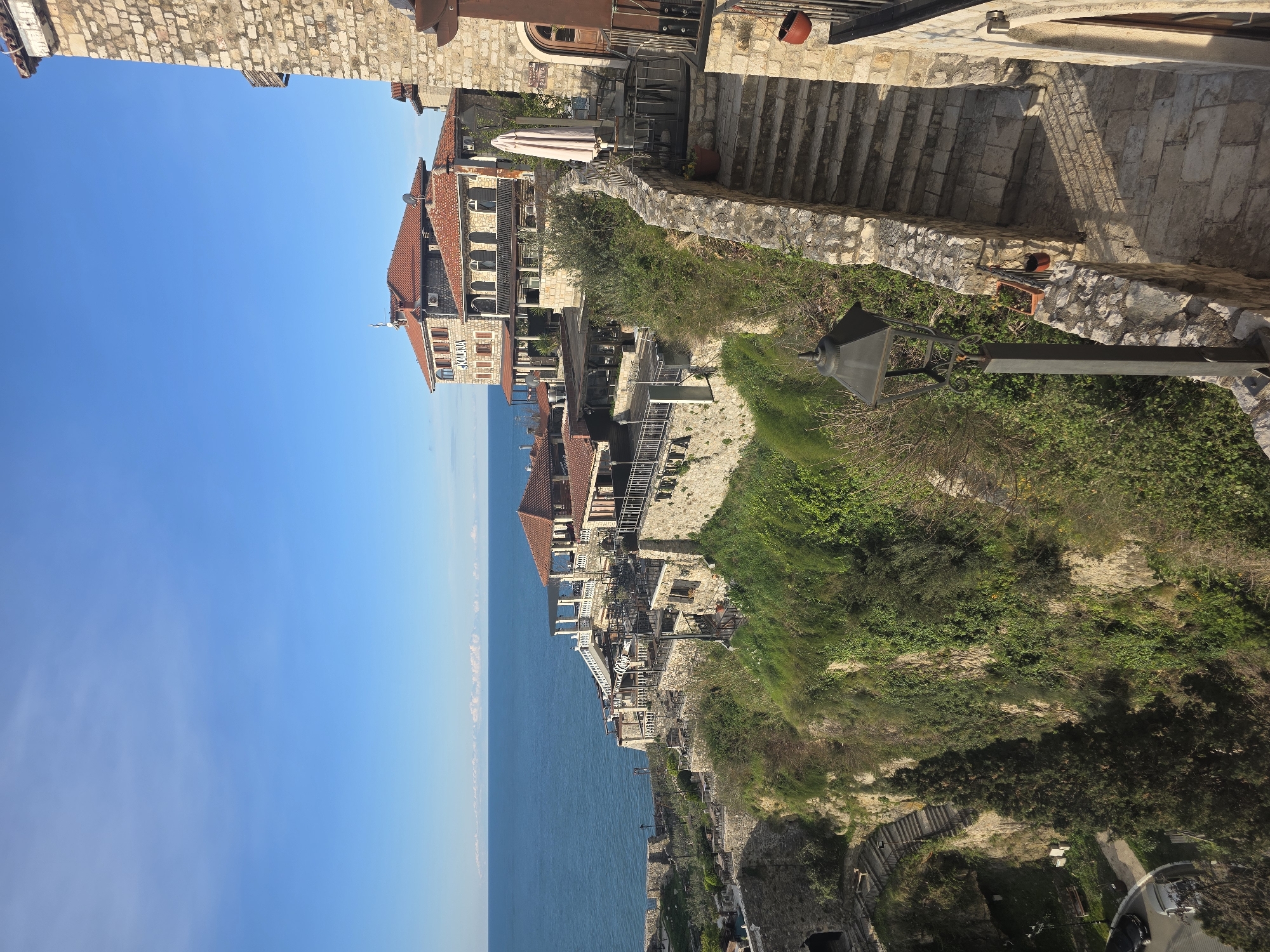 The image shows a coastal view in Ulcinj, Montenegro. On the left is the Adriatic Sea, with a clear blue sky above. Buildings with red-tiled roofs are constructed along the cliffside, blending with the natural stone formations. A series of stone staircases and pathways wind through lush green vegetation growing on the slopes. A vintage-style lamppost is visible on the right side of the path. The upper right corner shows architectural stone details and more buildings.