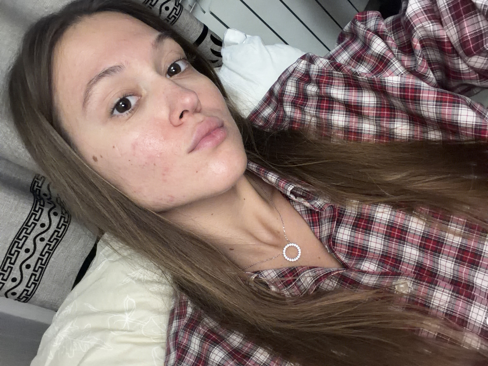 A person lies on a bed. The person has light brown hair, brown eyes, and fair skin. The person wears a red and white plaid shirt and a silver necklace. The location is inside a room with a bed and a pillow. The photo does not provide enough detail to identify the city as Nakhodka, Russia.
