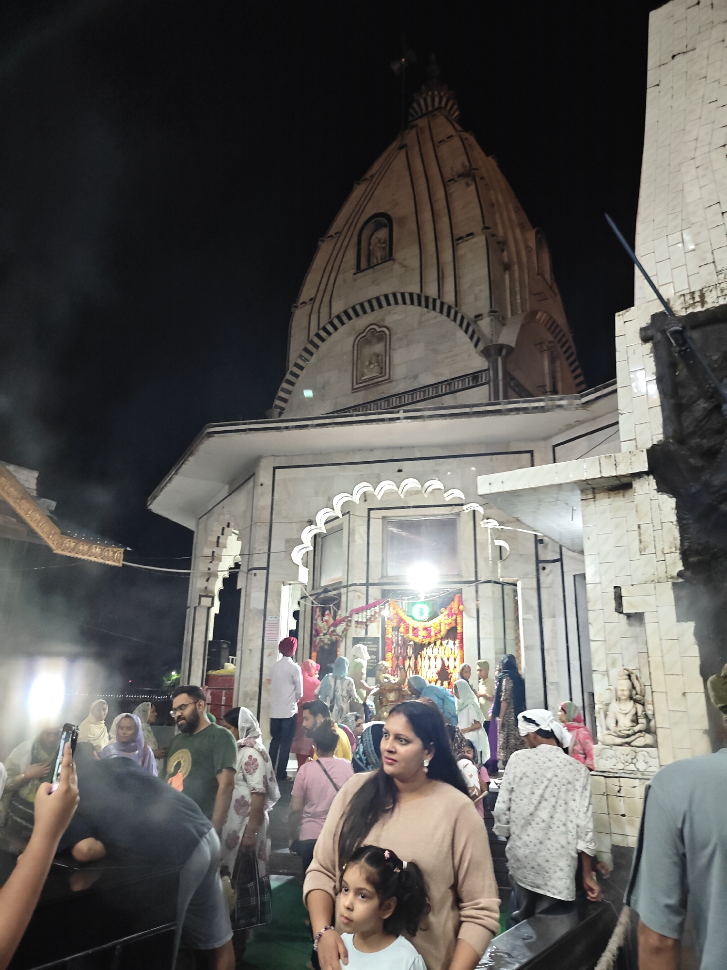 The image captures a vibrant night scene outside a Hindu temple in Kasol, India. The central subject is the temple, a multi-story structure constructed from light-colored stone, possibly marble. It features a distinctive, ribbed dome (shikhara) tapering towards the sky, with arched windows and decorative patterns adorning its facade. The main entrance area is brightly illuminated, revealing an ornate archway and what appears to be a colorful shrine or altar inside.
A large number of people are gathered in the foreground and mid-ground, suggesting a religious event, worship, or festive occasion. The crowd consists of men, women, and children of various ages, dressed in a mix of traditional Indian attire, such as kurtas, sarees, and head coverings, alongside more modern clothing. Many individuals are moving, with some appearing to enter or exit the temple, while others are standing and observing. In the immediate foreground, a woman in a light brown top holds a young girl, both looking towards the left. To their left, another person is seen holding a smartphone, seemingly taking a photo.
The time of day is unmistakably night, with the sky appearing pitch black above the well-lit temple structure. Artificial lights create bright glows around the temple entrance and on the left side of the frame, contributing to a lively atmosphere. A hazy or smoky effect is visible in the air, particularly on the left, possibly from incense or other rituals. On the right side, near the temple entrance, a stone statue, resembling Lord Shiva meditating, is clearly visible. The overall mood is one of devotion and community gathering. No clearly legible text is visible within the image.