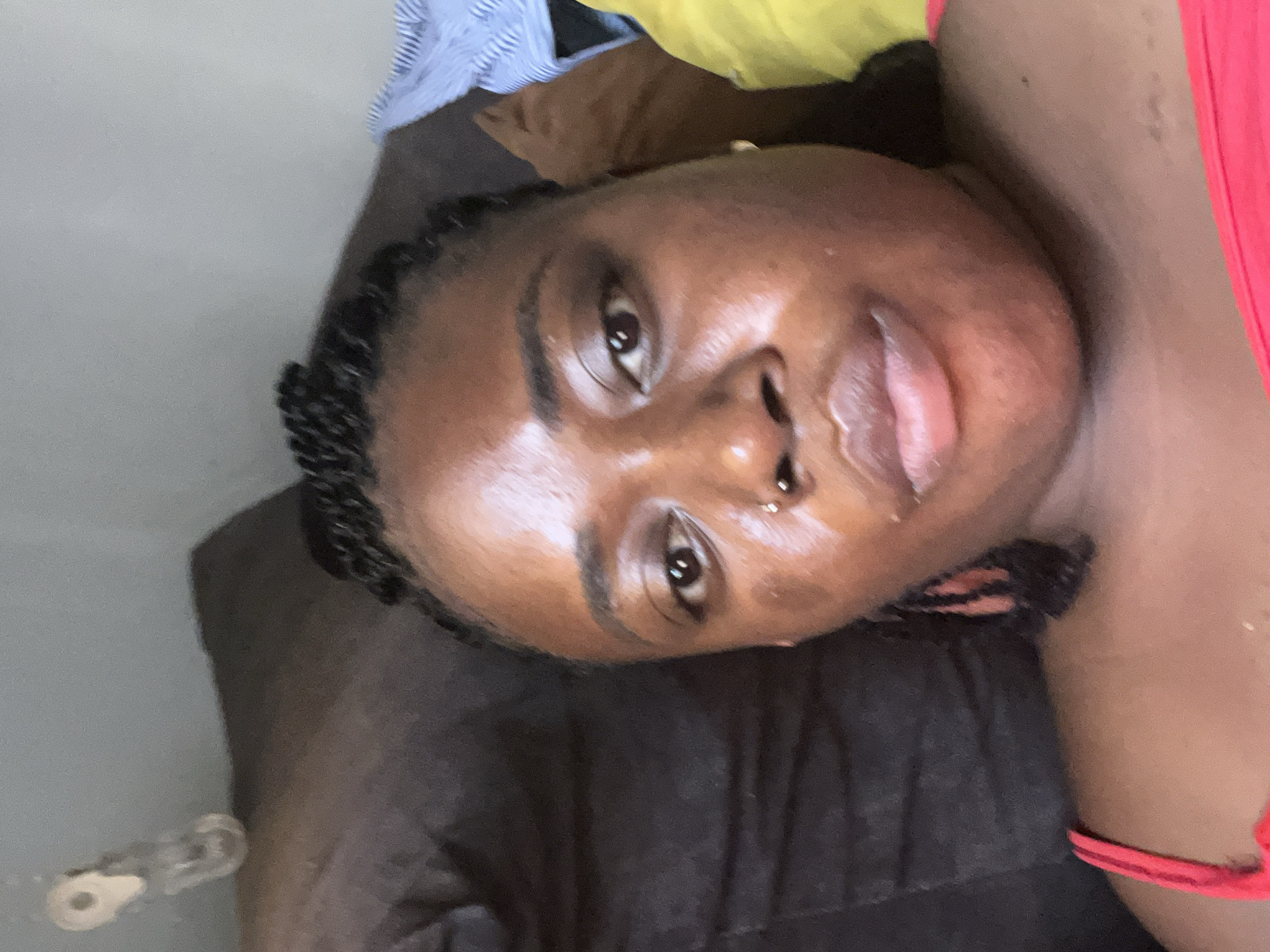 The image is a close-up selfie of a young Black woman with braided hair, a nose ring, and wearing a red top. She appears to be lying down on a brown surface, possibly a couch or bed. In the background, a striped blue and white fabric and a yellow fabric are visible, suggesting a domestic or casual setting. The lighting suggests it is daytime, possibly indoors. The background also shows a blurred object that resembles a key or a small hook attached to a wall. The woman's skin appears to have a dewy or oily sheen, and there are some blemishes. The image's focus is on the woman's face and upper body. There is no specific identifiable location within Abuja, Nigeria.