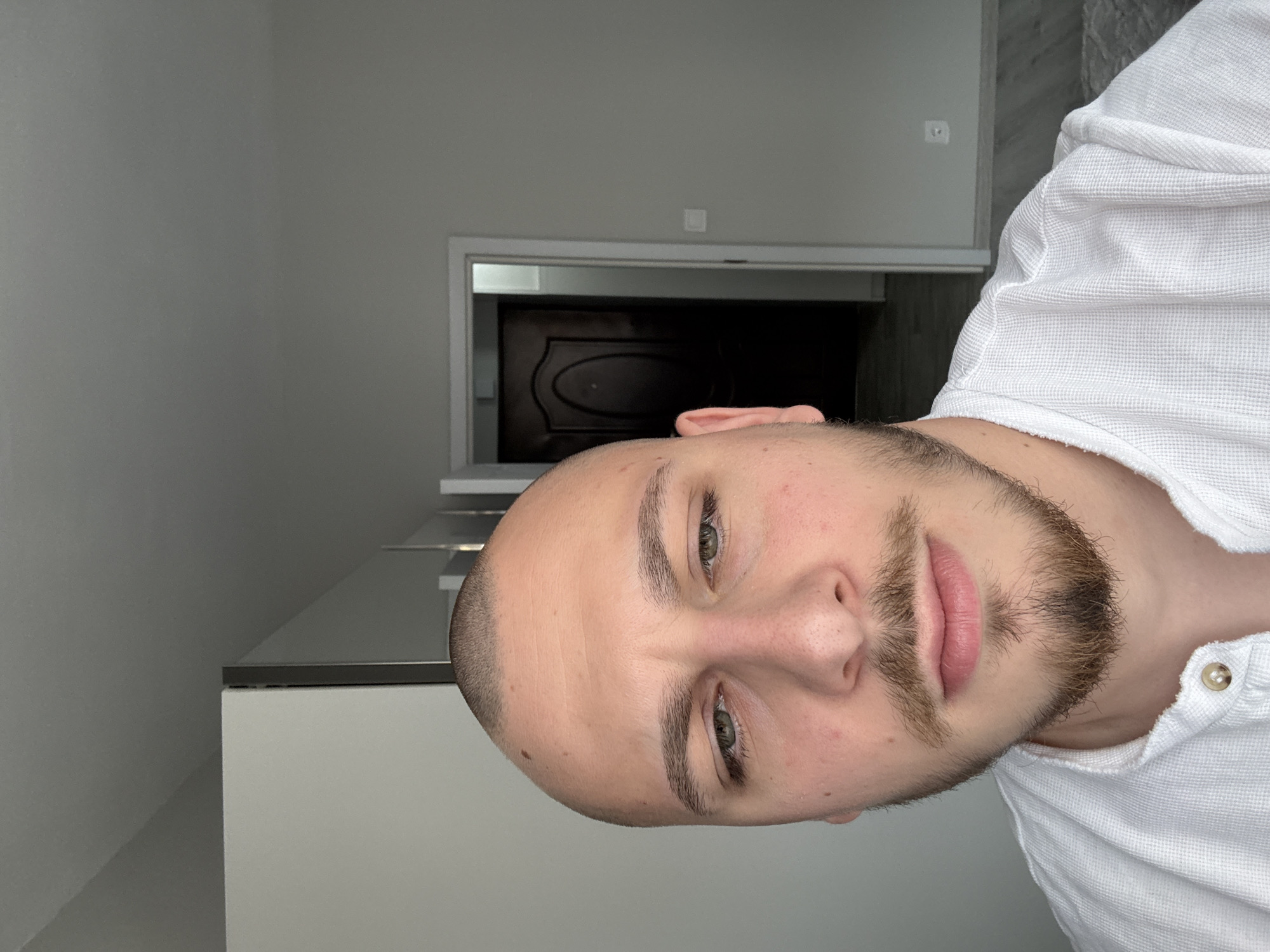 A light-skinned adult male with a shaved head, light brown mustache, and goatee is centered in a selfie-style photograph. He wears a white, textured Henley shirt with two visible light-colored buttons. His greenish-hazel eyes are slightly narrowed, directed at the camera. The setting is an indoor residential space located in Stara Zagora, Bulgaria. The background features plain light grey walls and ceiling. To the subject's right, an open doorway with white trim reveals a dark brown wooden door with an arched panel design. A white electrical outlet is visible on the wall to the right of the doorway. To the subject's left, a light-colored furniture unit, possibly a wardrobe or cabinet, with a dark vertical trim is partially visible. The lower right foreground shows a light grey wood-look floor partially covered by a light-colored, patterned rug. The scene is lit by natural light originating from the left.