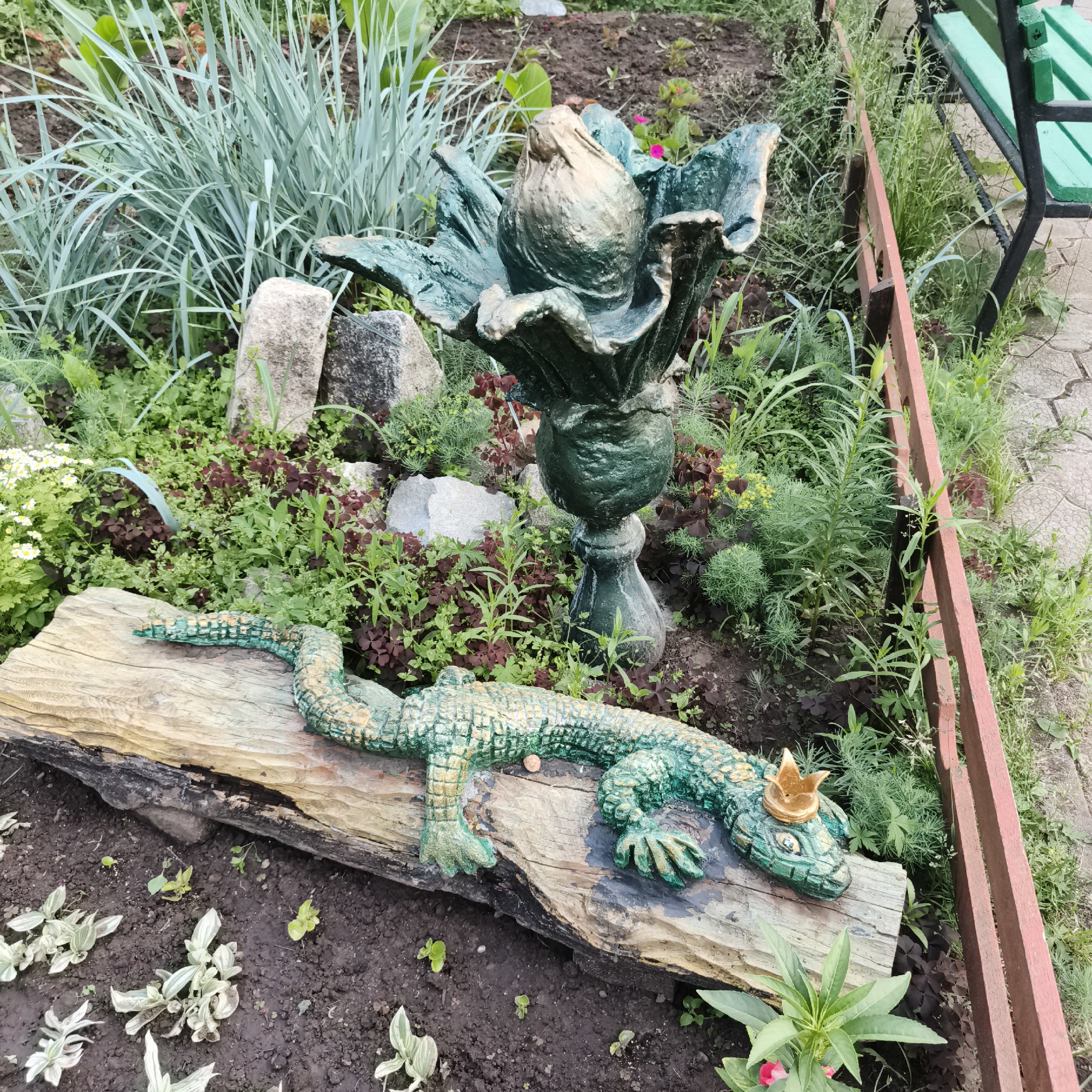 This is a high-angle, medium shot of a garden in the city of Chaikovsky, Russia. The garden features a concrete sculpture of a green alligator wearing a gold crown, lying on a log. Behind the alligator is a large, dark green, flower-shaped sculpture on a pedestal. The ground is covered with soil, small plants, and scattered rocks. In the background, there is a wooden fence and a green metal bench. The overall impression is of a decorative garden with whimsical elements.