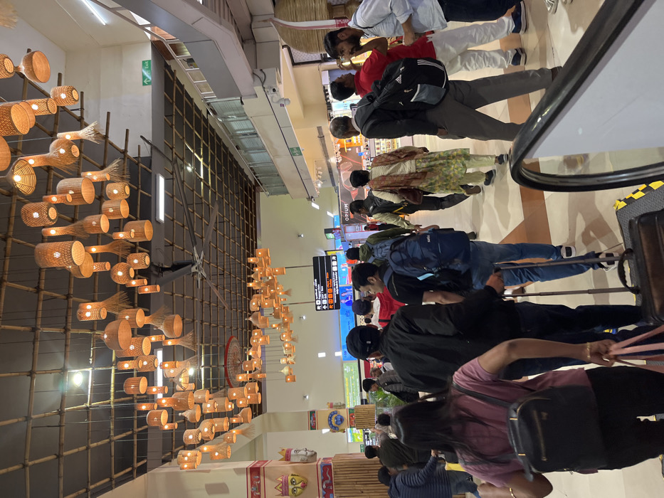 This media file captures an indoor scene, likely within an airport terminal or a large public building in Guwahati, India, given the unique local decorative elements. The image is taken from a slightly elevated perspective, looking across a spacious hall where a queue of people is forming.
The primary subjects are numerous individuals, predominantly adults, standing in a line extending towards the right side of the frame. Many are carrying personal bags, backpacks, and some have luggage, including a large black suitcase visible in the foreground on the right, next to what appears to be an escalator handrail. The people are dressed in casual attire, with a mix of traditional and modern clothing visible, such as a person wearing a patterned kurta. Most faces are partially obscured or seen from behind as they queue.
The setting is characterized by a distinctive and elaborate lighting installation made from bamboo and woven materials, suspended from the ceiling. These fixtures, resembling baskets or fish traps, emit a warm, golden light, creating a decorative and culturally rich ambiance. Similar bamboo lattice work adorns the walls. In the background, various storefronts and information signs are visible. One shop clearly displays the name "OPIUM". Another sign shows arrows and symbols, typical of public navigation. A green "EXIT" sign with a running man icon is also present high on a wall.
The activity taking place is primarily queuing, suggesting people are waiting for a service, possibly check-in, security, or boarding at an airport. The presence of luggage and the general environment strongly support this interpretation. The time of day is not explicitly clear, as the scene is artificially lit, but the warm glow from the lamps suggests an inviting atmosphere. There are no strong discernible emotions on the faces of the people, most appear to be waiting patiently. The overall impression is one of organized movement within a well-decorated, modern public space with traditional design influences.