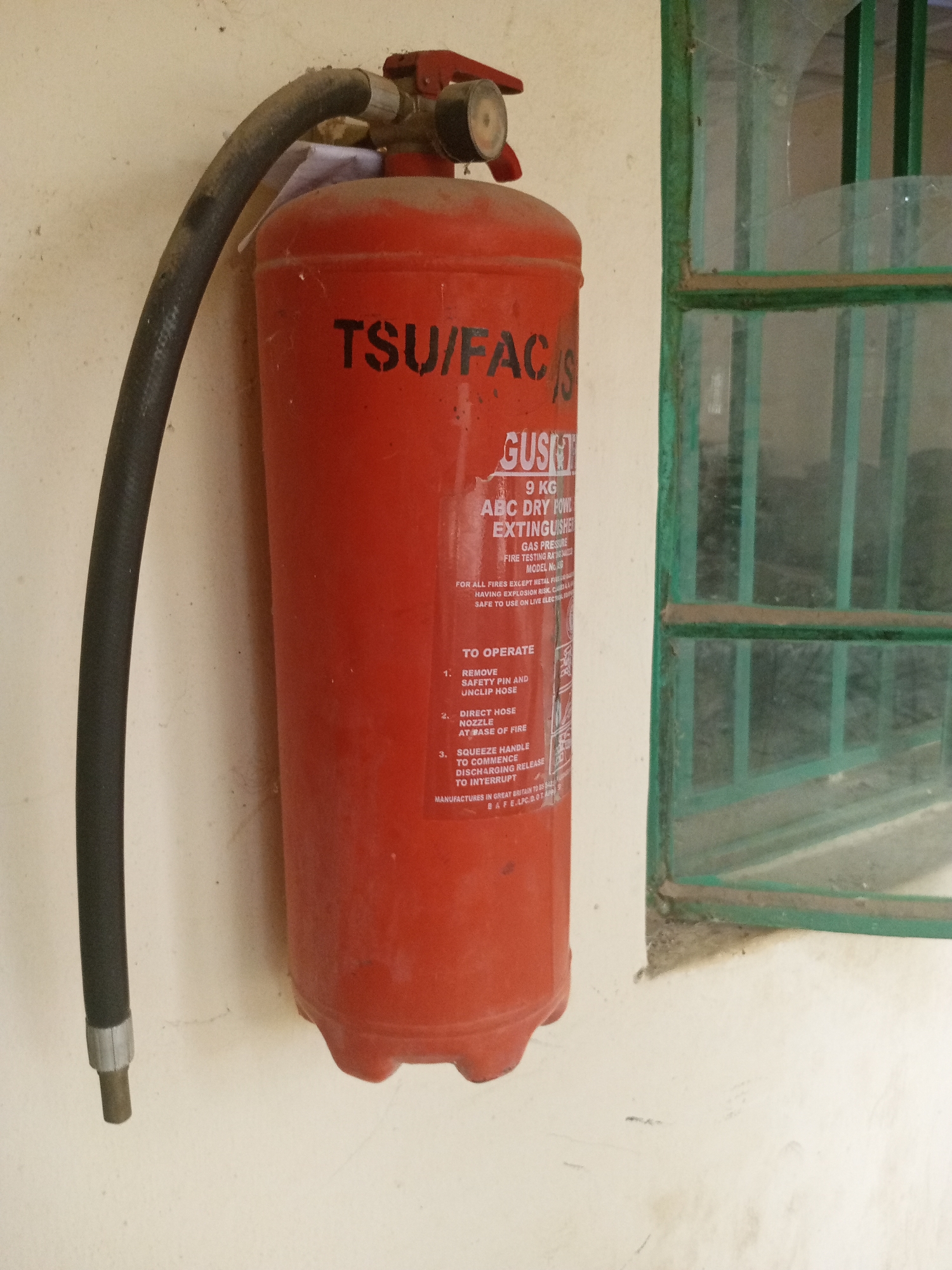 This image shows a red ABC dry powder fire extinguisher mounted on a textured cream-colored wall. A black, corrugated hose is attached to the top of the extinguisher. To the right of the extinguisher, part of a window with green bars is visible. The window pane appears slightly dirty and potentially cracked in one spot. The overall lighting suggests it might be daytime, but it's difficult to determine the exact time or weather conditions.
Text visible on the extinguisher includes "TSU/FACIS", "GUSN", "9 KG", "ABC DRY POWDER EXTINGUISHER", "GAS PRESSURE", "FIRE TESTING RA", and "MODEL No." It also provides operating instructions: "1. REMOVE SAFETY PIN AND UNCLIP HOSE", "2. DIRECT HOSE NOZZLE AT BASE OF FIRE", and "3. SQUEEZE HANDLE TO COMMENCE DISCHARGING RELEASE TO INTERRUPT". A line at the bottom reads "MANUFACTURES IN GREAT BRITAIN TO BS 5423".
The extinguisher appears to be functional but shows some signs of dust and age, consistent with being mounted in a practical, perhaps industrial or public, setting. The location context is Jalingo, Nigeria.