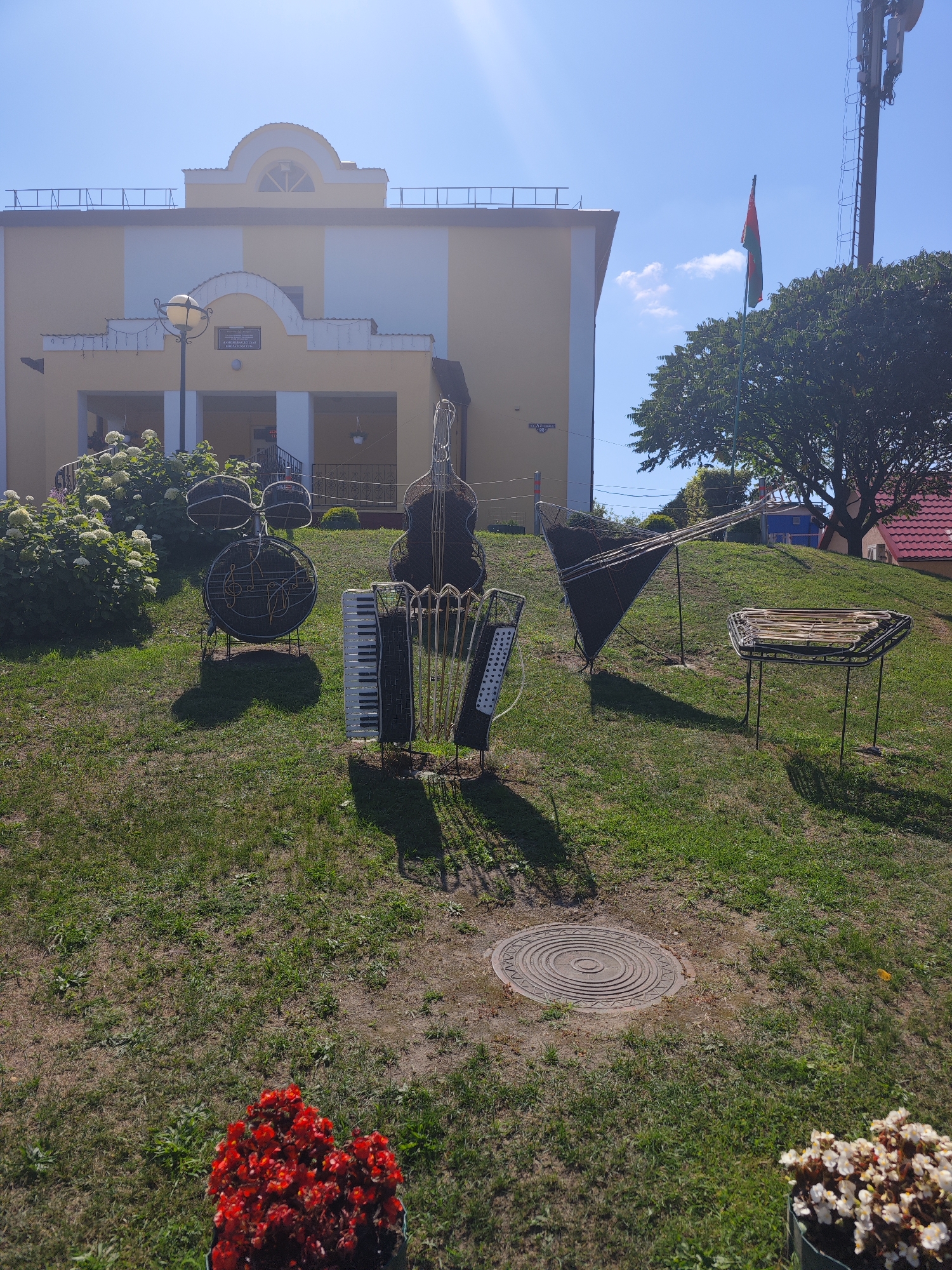 In Kamyanyets, Belarus, on a bright sunny day, a grassy hill slopes upwards towards a light yellow and white building with a curved architectural detail at the top. In the foreground, a collection of wire sculptures depicting musical instruments is arranged on the lawn. These sculptures include a drum set with musical notes, an upright bass, and an accordion. Further up the hill, there is a triangular stringed instrument sculpture and a small table-like sculpture. A tall telecommunications tower with a flag attached to it stands to the right of the building. The environment is a public space with manicured grass, bushes, and two pots of flowers, one with red flowers and another with white flowers, visible in the bottom corners of the image. A manhole cover is also visible on the grass. The scene captures an outdoor art installation with a backdrop of a building and urban infrastructure.