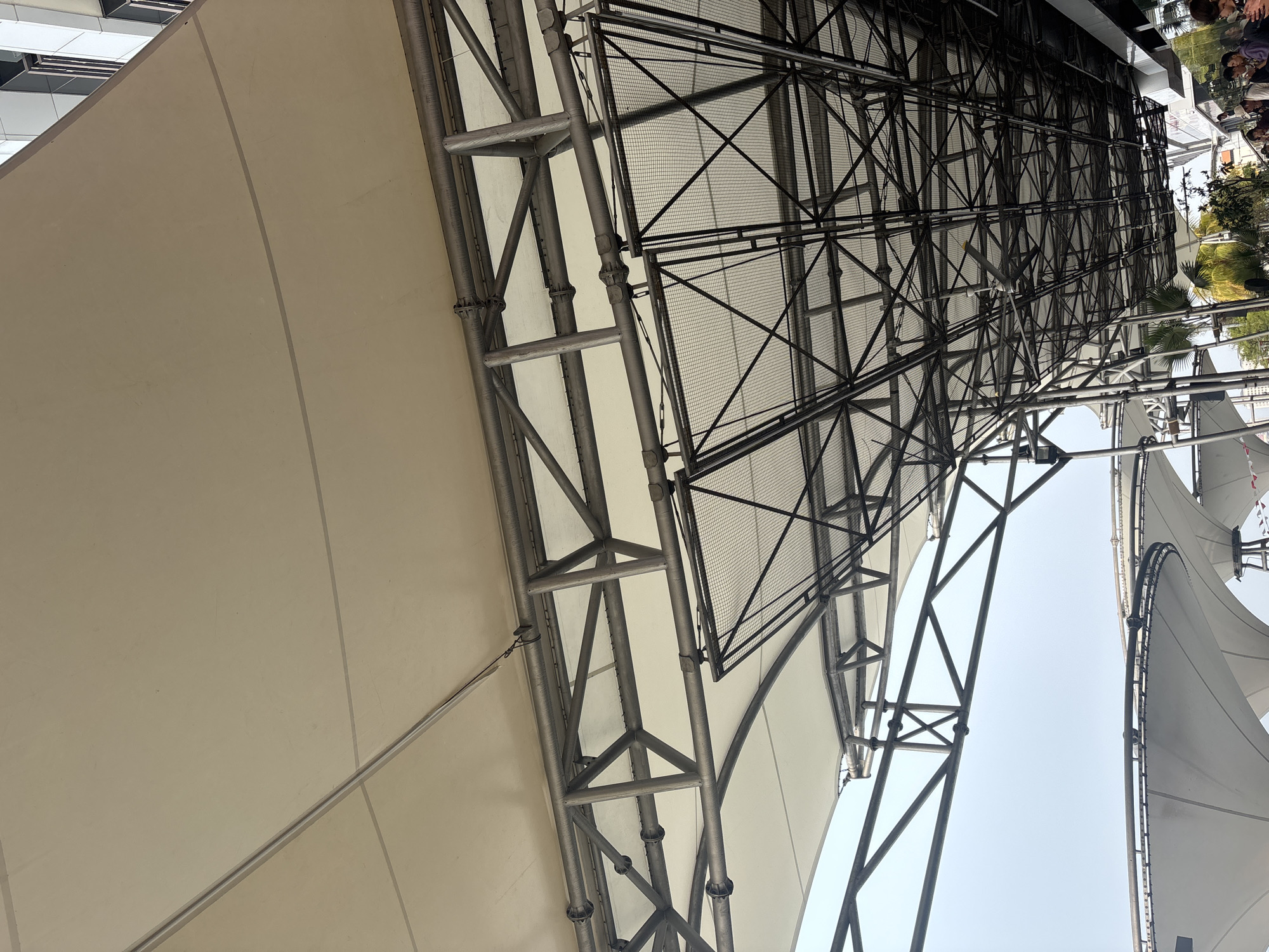 The image shows an overhead view of a canopy structure with a metal framework supporting a beige fabric covering. Below the fabric, a complex network of black metal beams and mesh forms a ceiling, with a ceiling fan visible. The scene appears to be outdoors, possibly a public space or a venue. In the lower right corner, a few people are visible, some of whom appear to be looking at their phones. To the right, part of a modern building with windows is visible. Palm trees are present in the background on the left. The sky above is overcast. The structure suggests it is located in an urban setting in North Jakarta, Indonesia, likely a covered walkway or seating area.