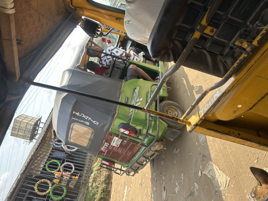 The image depicts a street scene in Umuode, Nigeria, viewed from inside a yellow tricycle. In the foreground, parts of the observer's yellow tricycle are visible, including a rusty yellow metal frame and a black textured seat. A brown animal ear is partially visible in the bottom left.
The central subject is a green TVS Keke Deluxe tricycle (auto-rickshaw) positioned on an unpaved dirt road. The tricycle has a dark grey canopy and features "TVS" on the rear and "DELUXE" and "BOXER" text on its side. Its rear bumper is a metal rack, with brown material (likely mud or dirt) adhered to the lower section. A Nigerian license plate, partially obscured, is visible on the rear. Inside the green tricycle, two individuals are seated: one wearing a red shirt and another wearing a black and white patterned shirt. The leg of a third person, wearing shorts, is also visible.
In the background, on the left, a single-story commercial building with a corrugated metal roof is present. Its facade features vertical metal bars, and several colorful ring-shaped objects (yellow, green, white, pink) are displayed on the exterior, possibly merchandise. Above the building, against a clear sky, an elevated rectangular water tank and utility wires are visible. The ground is a light brown dirt road, scattered with small pieces of white debris. The scene is illuminated by strong daylight.