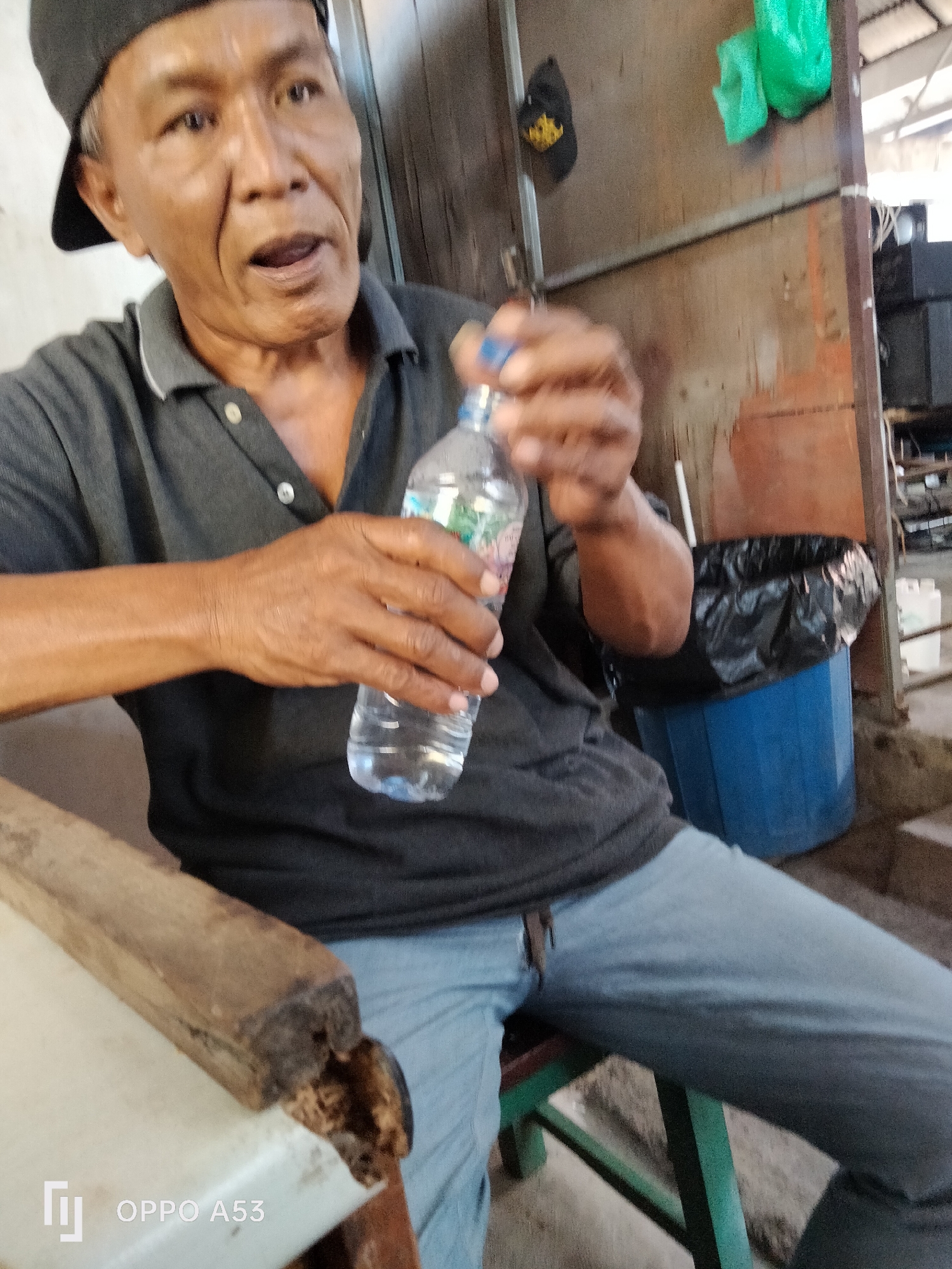 An Asian male wearing a black baseball cap and a dark grey polo shirt sits on a green metal chair. He is holding a clear plastic water bottle with a blue cap in his right hand and appears to be opening it or drinking from it. His left hand is near the bottle, possibly steadying it. The background consists of a wooden wall, a blue plastic bin with a black trash bag, and some shelves with what appear to be electronic equipment. A wooden table is in the foreground. The overall setting appears to be a utilitarian space, possibly outdoors or in an open-air structure, with elements suggestive of a workshop or informal market area. The image was captured in Denpasar, Indonesia.