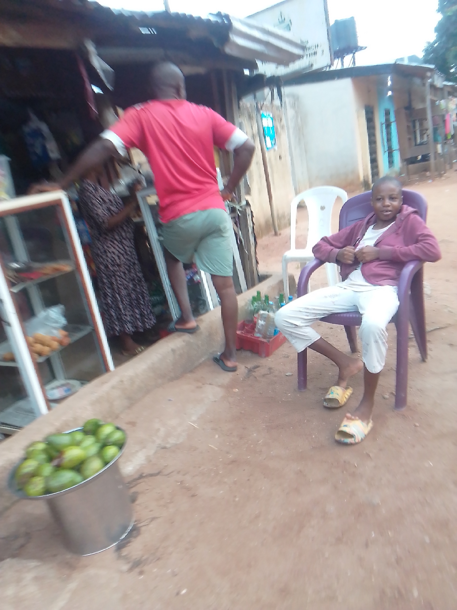 Here is a forensic description of the scene:
**Location:** Owerri, Nigeria. The scene appears to be in a semi-urban or roadside location.
**Subjects:**
* A young individual is seated in a purple plastic chair wearing light-colored pants, a white shirt, a maroon jacket, and slides.
* A man in a red shirt and green shorts stands near a shop.
* Another individual in a long printed dress stands near the shop.
**Objects:**
* A metal bucket filled with green fruits.
* A glass-fronted display case exhibiting food items is located at the shop.
* A red plastic crate.
* Plastic bottles.
* Two plastic chairs (purple and white).
* A building with a corrugated metal roof and various structural elements.
* A building with a corrugated metal roof and a water tank on top of it.
**Actions and Interactions:**
* The seated person is positioned in a chair in front of the shop, facing the camera.
* The man in the red shirt appears to be interacting or associated with the shop.
**Environment:**
* The ground is a bare earth surface.
* The background includes a mix of building structures and what appears to be a street or roadside environment.
* Evidence of a small commercial establishment/shop is seen.