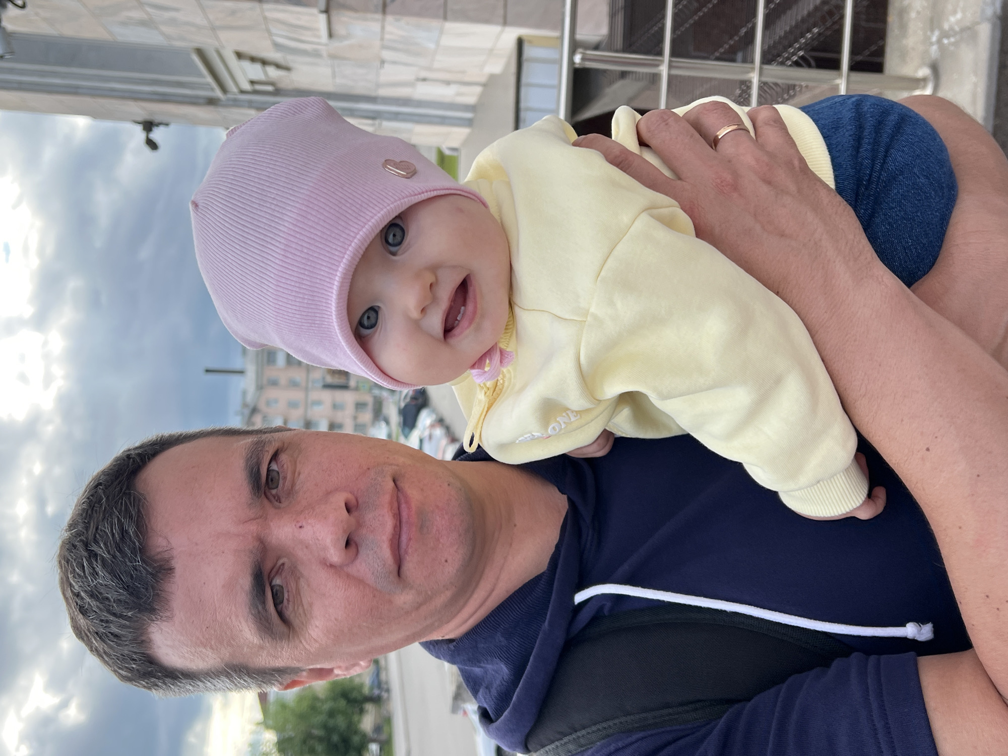 An adult male with short, graying brown hair and brown eyes, wearing a navy blue hooded sweatshirt, holds a baby girl. The baby has blue eyes and wears a pink knitted hat and a light yellow hooded jacket. The scene is outdoors, with a building and parked cars visible in the background. The location is on a street in Izhevsk, Russia, likely near a residential area given the building style and parked vehicles. The man is looking at the camera, while the baby is looking slightly to the side and smiling. The interaction suggests a parent and child moment.